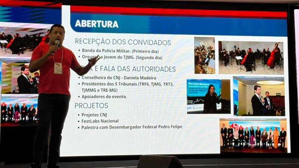 Um homem de camiseta e calça escura está em um palco, falando em um microfone. Atrás dele, uma tela grande exibe um slide.