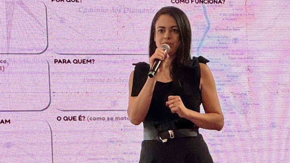 Uma mulher de vestido escuro, com um cinto e mangas bufantes discretas, está no palco segurando um microfone e falando. Atrás dela, uma tela exibe um slide.