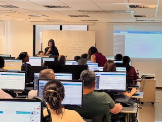A imagem mostra um ambiente interno de sala de aula ou treinamento com tecnologia. Há um grande número de pessoas sentadas em mesas individuais ou duplas, voltadas para monitores de computador.

A imagem apresenta uma mulher de pé na frente da sala, próxima a um quadro branco com escrita e um datashow (projetor) que exibe uma tela de computador. Os participantes estão sentados em frente aos seus computadores, alguns olhando para a frente.