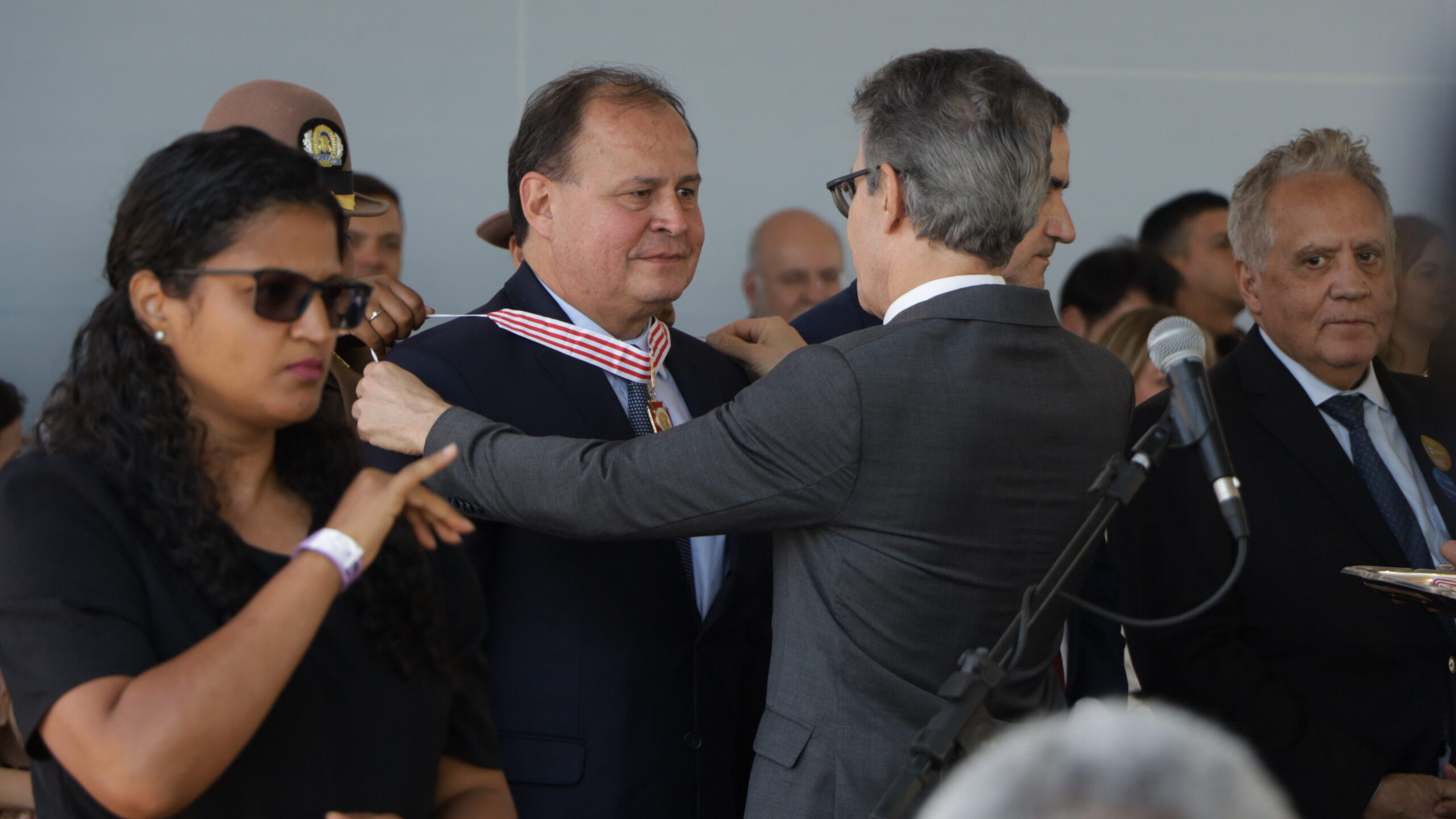 Um homem de terno cinza coloca uma medalha com fita vermelha e branca no pescoço de um homem de terno azul-marinho durante uma cerimônia.