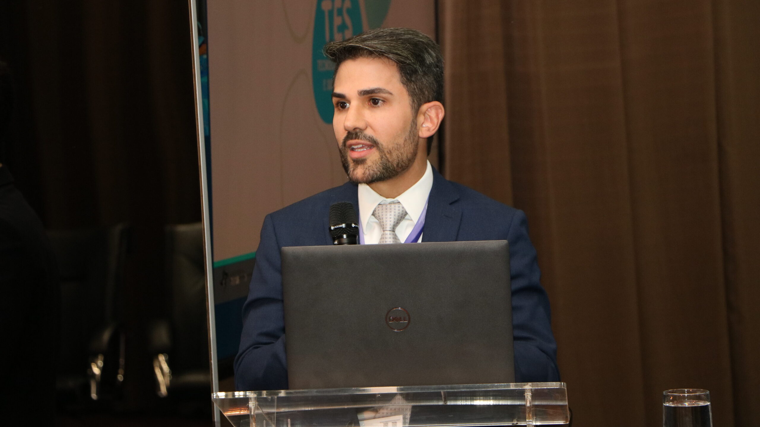 Foto em plano médio de um homem jovem, com barba e cabelo escuro, vestido com um terno azul-marinho, camisa branca e gravata clara, discursando em um evento.

Ele está em pé atrás de um púlpito de acrílico, com um notebook preto aberto à sua frente. Ele segura um microfone com a mão direita, olhando para a frente.

O fundo é uma cortina marrom escura.