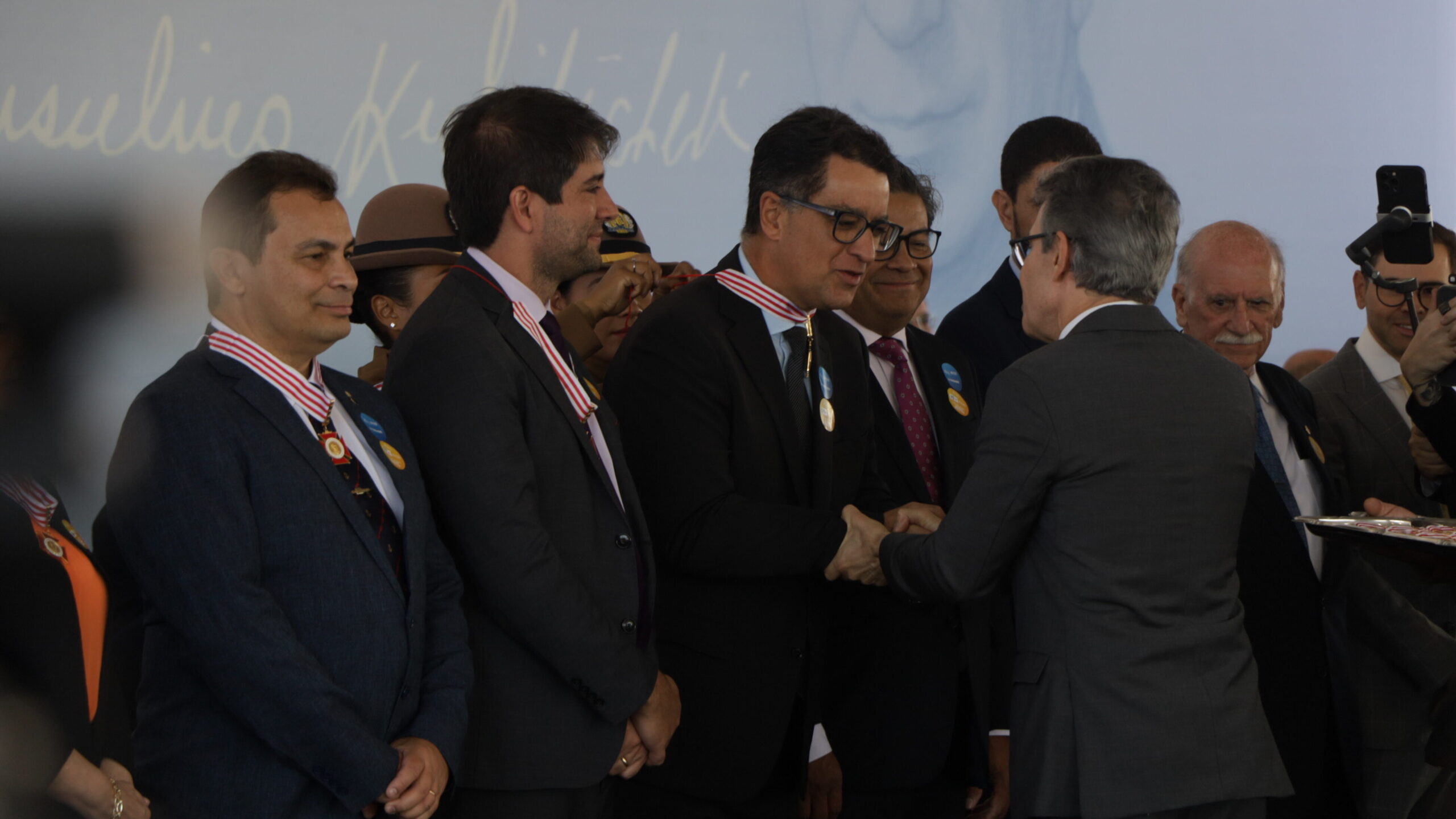 Dois homens de terno se cumprimentam com um aperto de mãos durante uma cerimônia. Vários outros homens de terno e com medalhas estão ao redor.