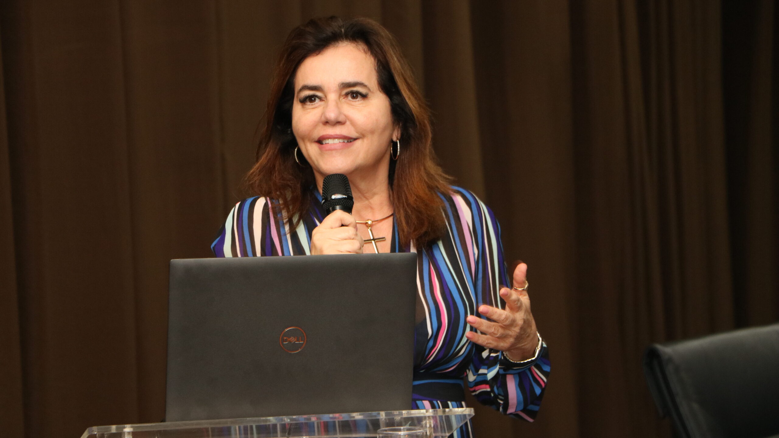 Foto de uma mulher falando em um evento.

Ela veste um vestido listrado e colorido (em tons de azul, roxo, rosa e preto), e usa um colar com pingente de cruz. A mulher está segurando um microfone com a mão direita e gesticula com a esquerda, sorrindo e olhando para frente.

À sua frente, há um notebook preto aberto sobre um púlpito de acrílico. O fundo é composto por uma cortina marrom escura.