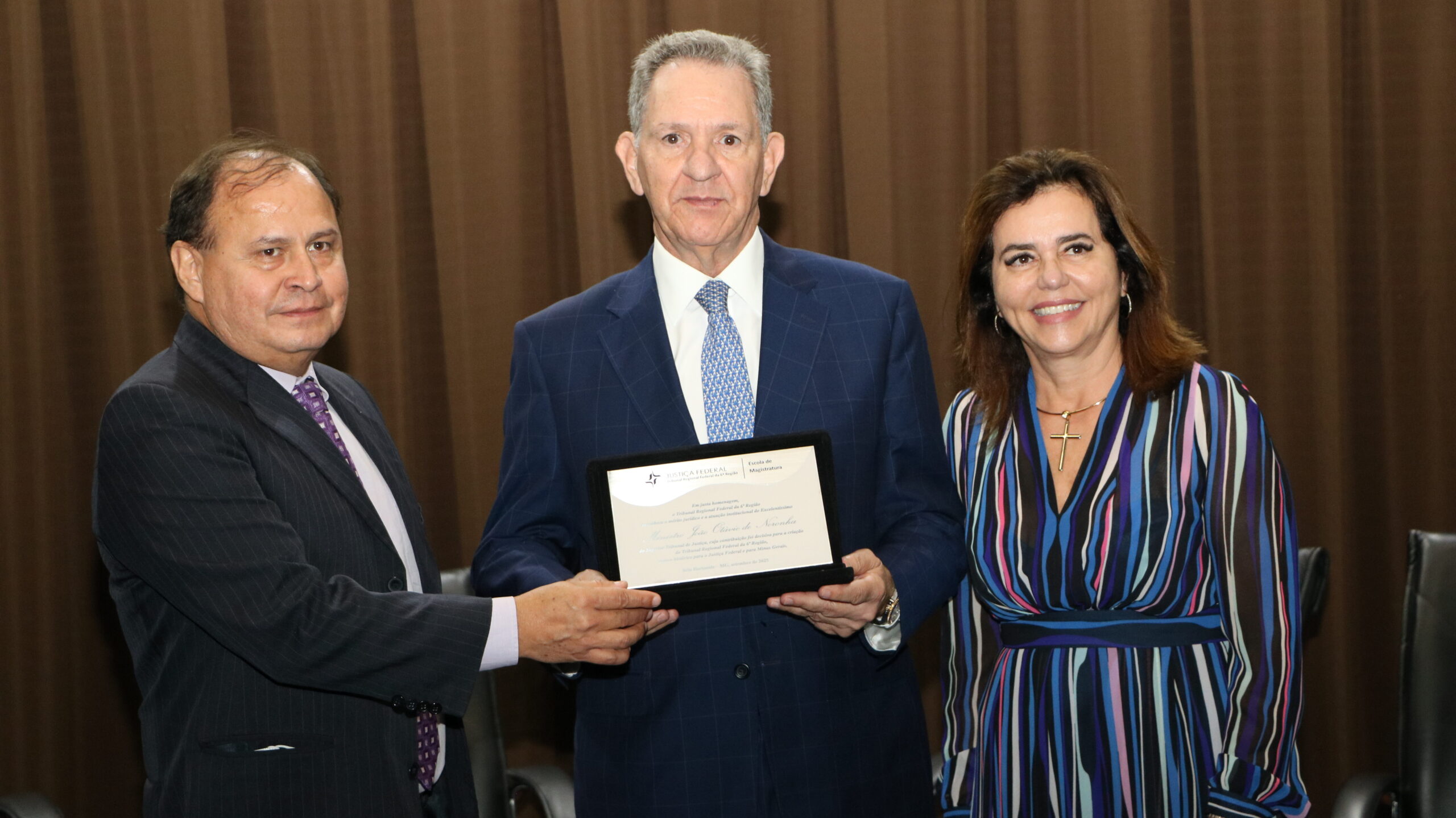 Foto institucional que registra um momento de homenagem ou entrega de certificado, mostrando três pessoas (dois homens e uma mulher) em pé, em primeiro plano.

O homem no centro, vestido com um terno azul-marinho e gravata de padrão azul claro, está segurando uma placa de homenagem/certificado preta.

O homem à esquerda (terno escuro e gravata roxa) e a mulher à direita (vestido listrado colorido em tons de azul, roxo e marrom) parecem estar participando da entrega e sorriem para a câmera.

O fundo é uma cortina marrom escura e há assentos pretos visíveis na parte inferior.