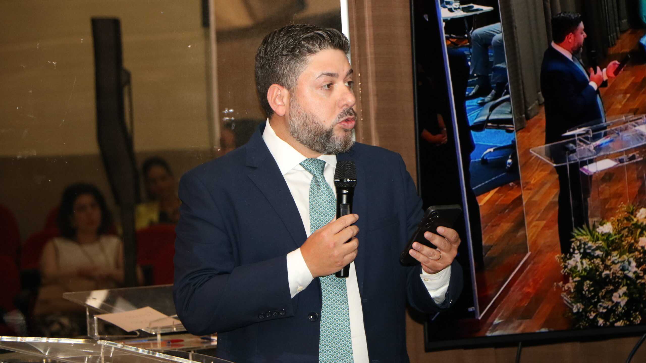 Foto de um homem barbado, vestido com um terno azul-marinho e gravata verde-claro/água estampada, falando em um evento.

Ele está segurando um microfone com a mão direita e um celular com a mão esquerda, olhando para a sua esquerda.

No fundo à esquerda, algumas pessoas estão sentadas e fora de foco.