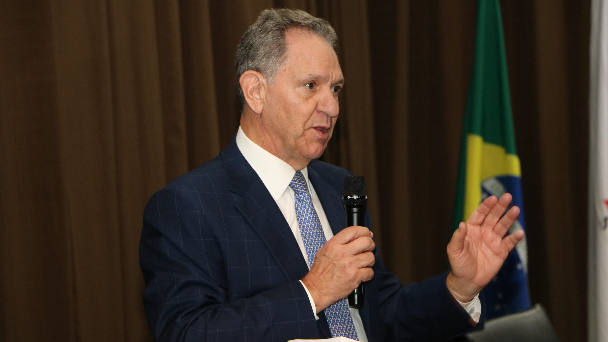Foto em plano médio de um homem de meia-idade, vestido com um terno azul-marinho e gravata estampada em tons de azul e branco, discursando em um evento.

Ele está segurando um microfone com a mão direita, enquanto gesticula com a mão esquerda aberta, olhando para a sua frente.

O fundo é composto por uma cortina marrom escura e, no canto superior direito, é visível parte de uma bandeira do Brasil.