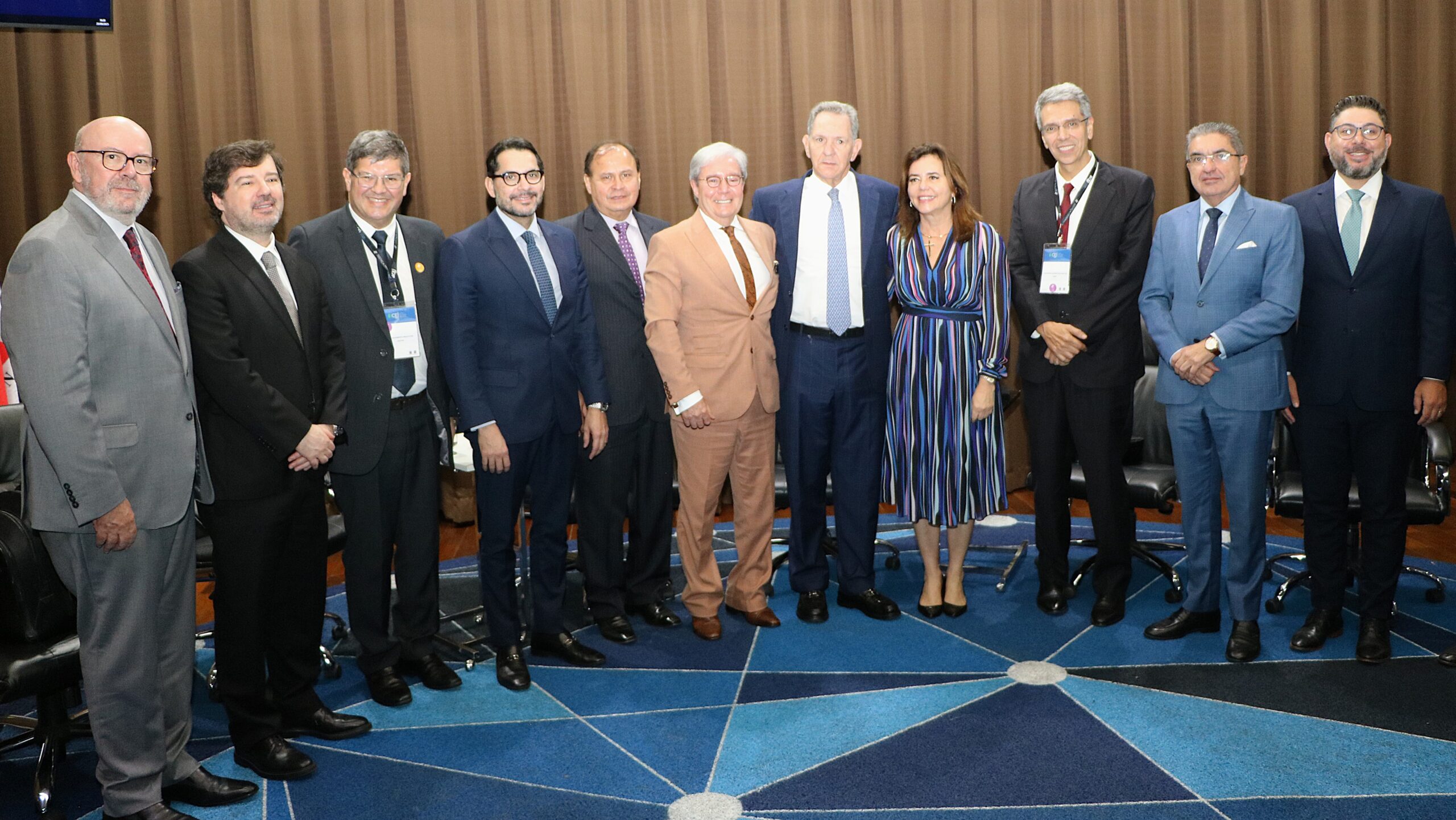 Foto de grupo em um evento corporativo ou institucional, mostrando onze pessoas (dez homens e uma mulher) em pé, lado a lado, posando para a câmera. O grupo está vestido com ternos sociais.

O fundo é composto por uma cortina marrom e o chão tem um tapete azul com um padrão geométrico de linhas brancas.