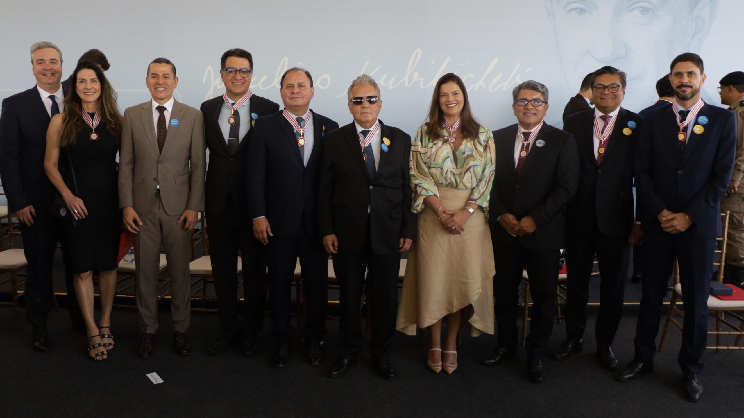 Grupo de dez pessoas, vestindo trajes formais (alguns com medalhas no pescoço), posam lado a lado em um evento solene com um painel com a imagem de Juscelino Kubitschek ao fundo.