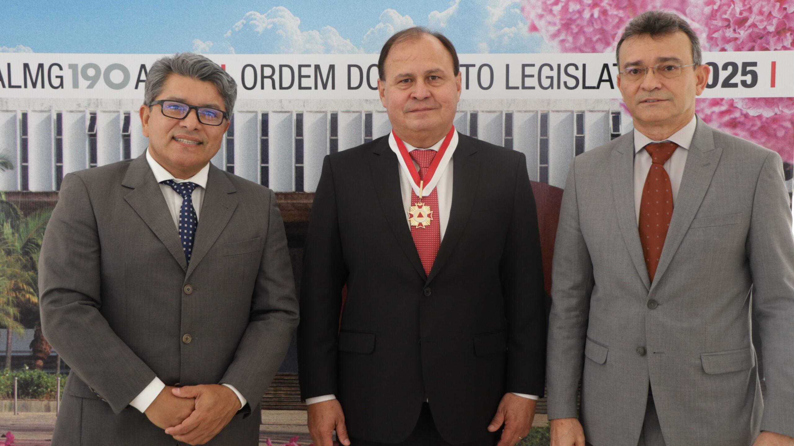 A imagem é uma fotografia que retrata três homens posando lado a lado em um evento formal. Todos vestem terno e gravata.