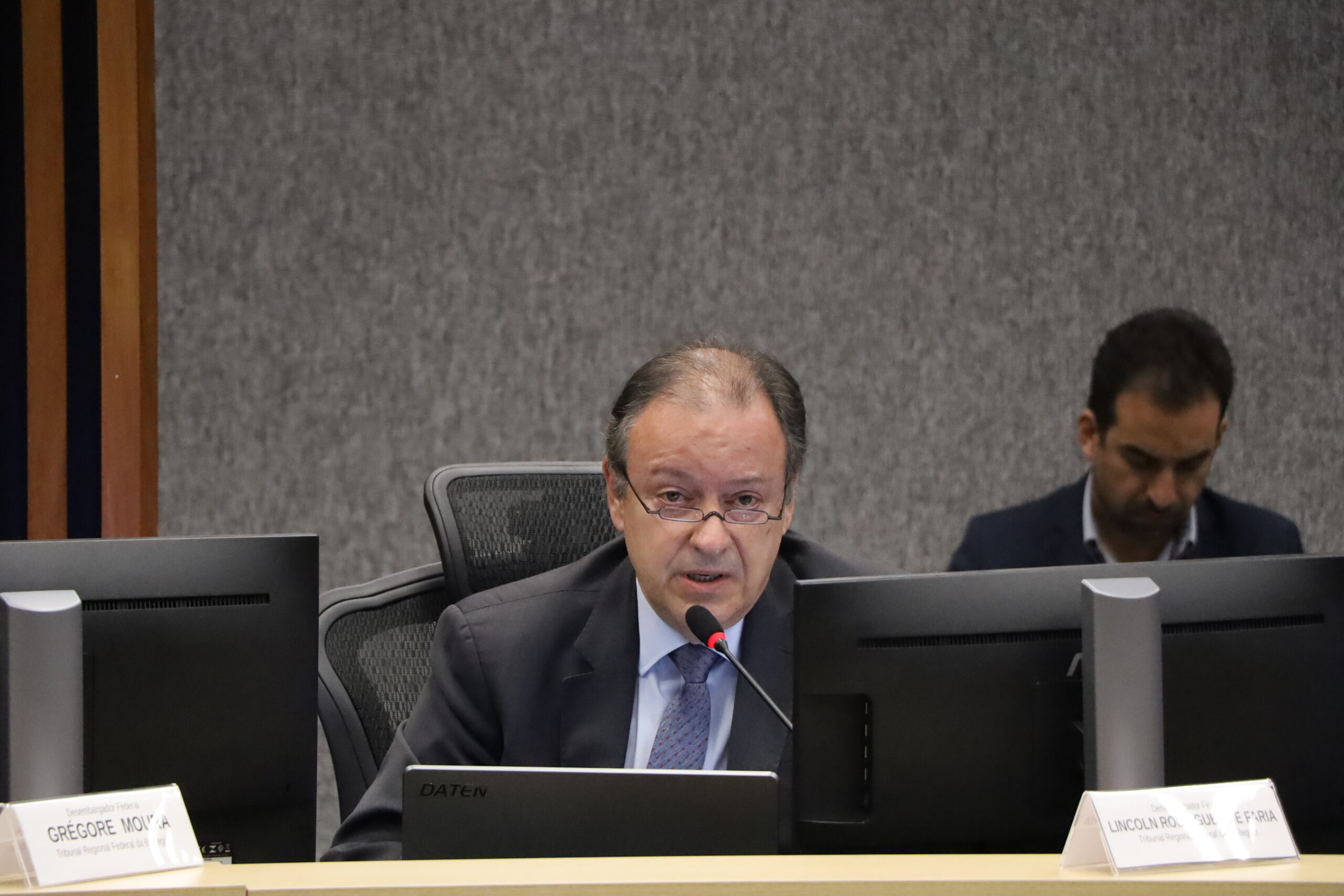 Um homem de meia-idade, vestindo terno escuro e gravata, sentado em uma mesa de reunião e falando em um microfone. Ele usa óculos e está olhando para a frente. Em frente a ele e ao seu lado, há monitores de computador. Outro homem, parcialmente visível, está sentado ao fundo, com a cabeça baixa. O fundo é uma parede de tecido cinza texturizado.