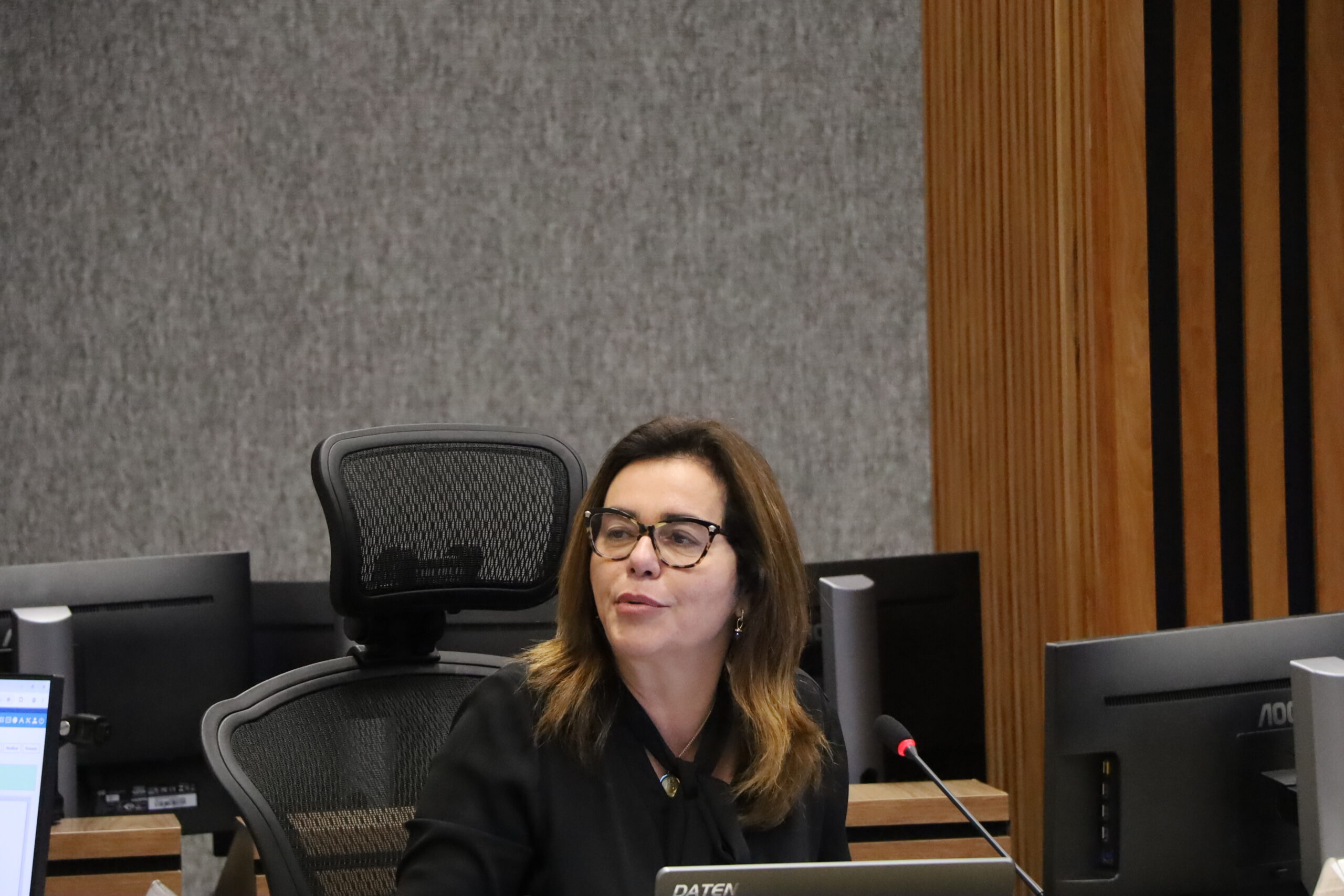 Uma mulher vestindo roupas escuras e óculos de armação clara, sentada em uma mesa de reunião e falando em um microfone. Ela está olhando para a frente. Há monitores de computador ao seu redor. O fundo é uma parede de tecido cinza texturizado.