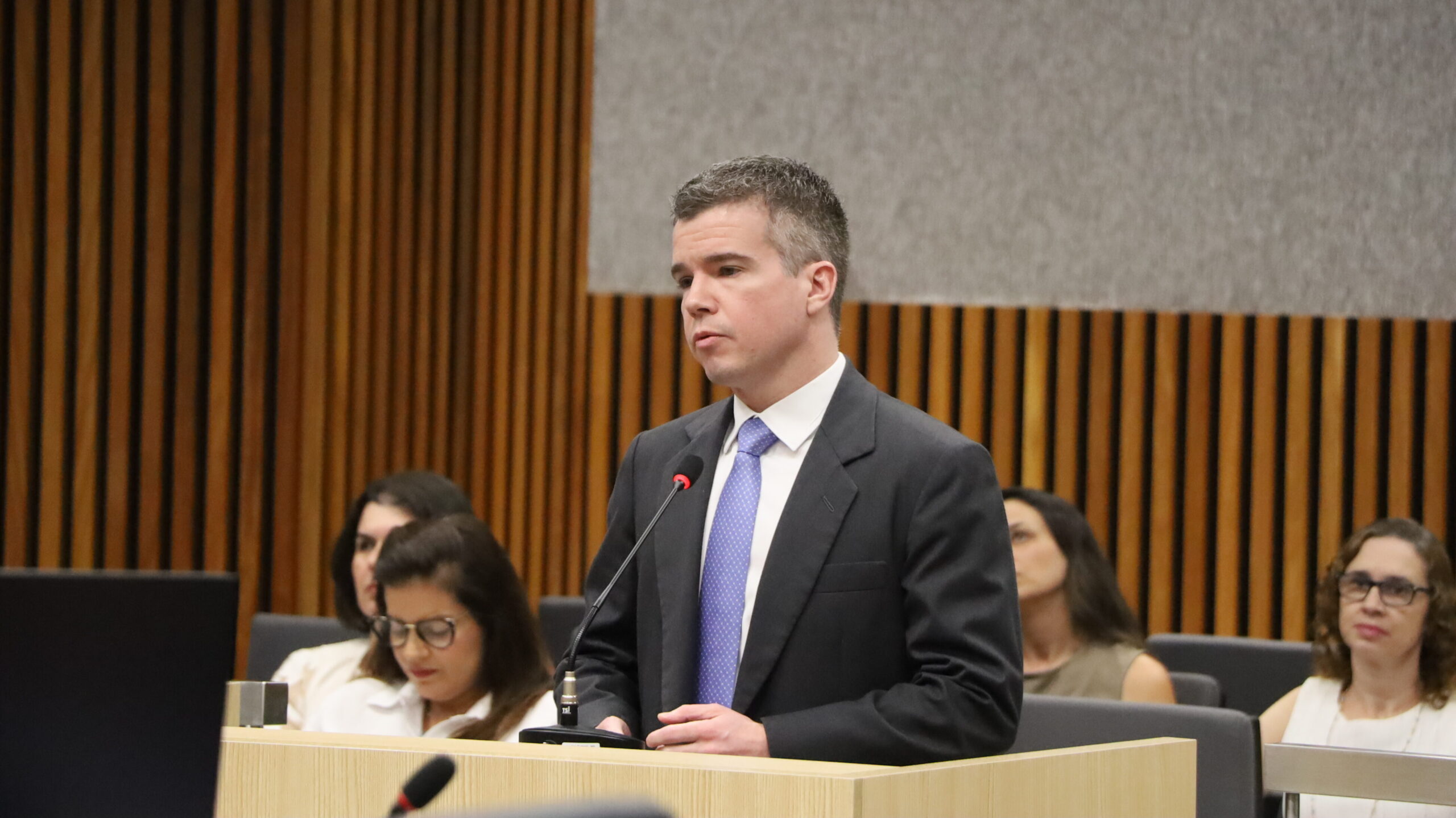 Um homem jovem, de cabelos curtos, vestindo um terno escuro e gravata azul, em pé atrás de uma mesa ou púlpito claro, falando em um microfone. Ele olha para a frente com uma expressão séria. Algumas pessoas estão sentadas atrás dele.
