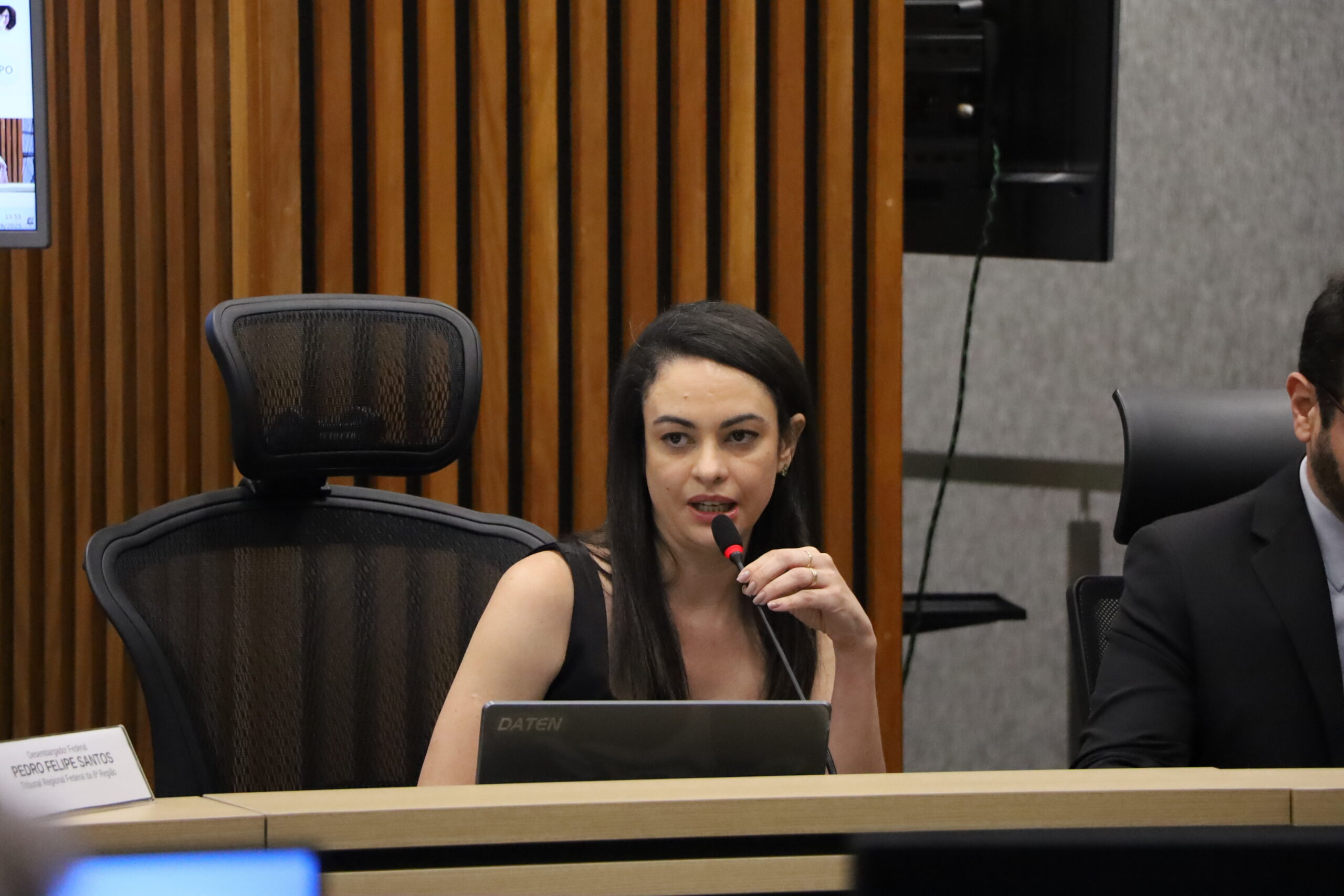 Uma mulher jovem, vestindo roupa escura sem mangas, sentada em uma mesa de reunião e falando em um microfone. Ela tem cabelos escuros e lisos. Em frente a ela, há um monitor de computador parcialmente visível.