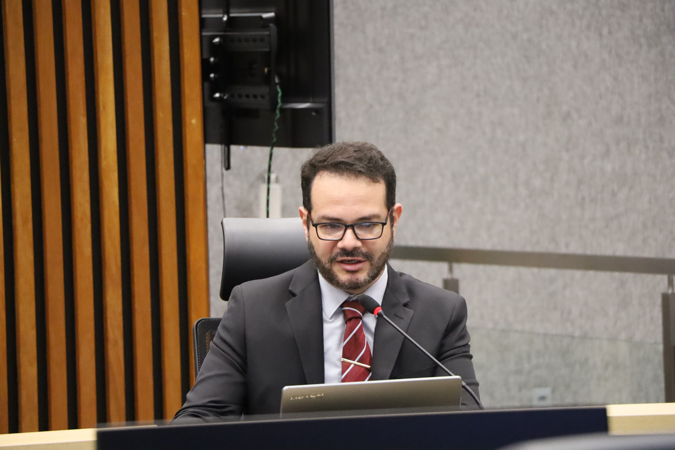Um homem barbudo, vestindo terno e gravata escura com listras diagonais claras, sentado em uma mesa de reunião e falando em um microfone. Ele usa óculos de armação escura. O fundo é uma parede de madeira ripada à esquerda e uma parede de tecido cinza texturizado no centro e à direita.