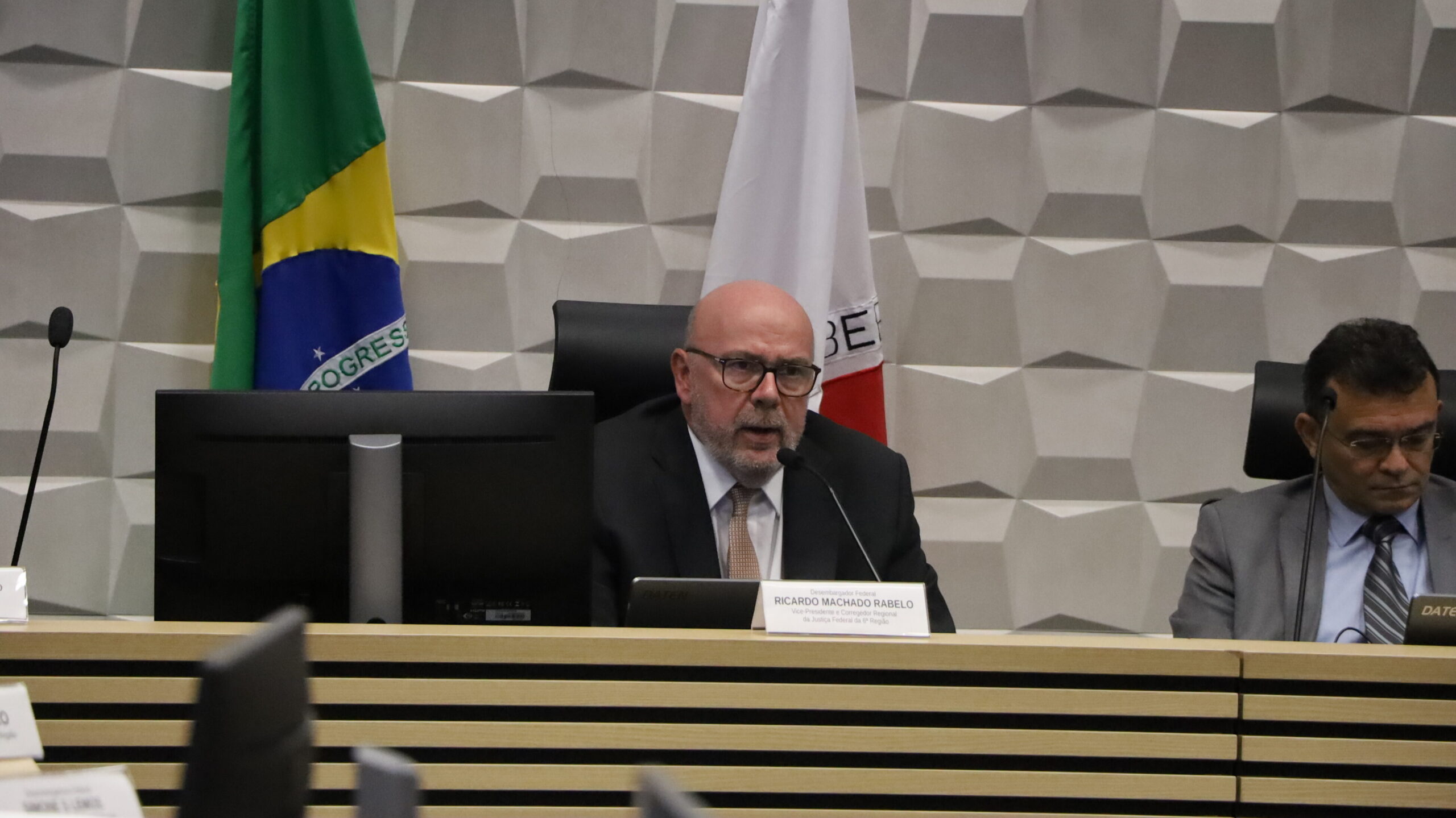 Um homem barbudo, vestindo terno e gravata, sentado em uma mesa de reunião e falando em um microfone. Ele usa óculos e está olhando para a frente. Em frente a ele, há uma placa de identificação. Um monitor de computador está posicionado à esquerda. Outro homem, parcialmente visível, está sentado à direita, olhando para baixo. Ao fundo, há uma parede com um painel em relevo branco e cinza, com as bandeiras do Brasil e de Minas Gerais visíveis.