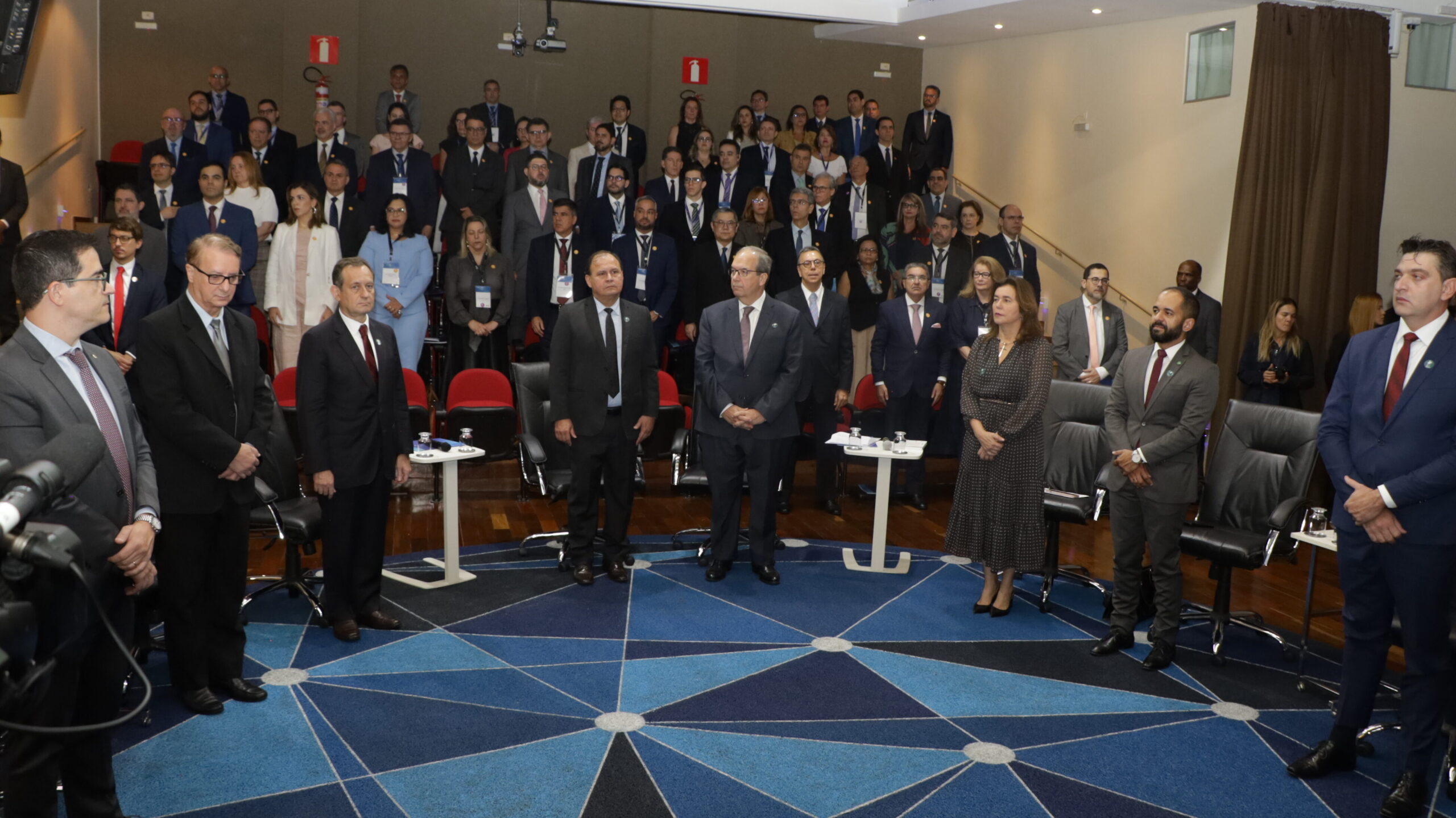 Uma foto de um grande grupo de pessoas em um salão ou auditório, todas em trajes sociais formais (ternos ou vestidos). O chão é coberto por um tapete azul com um padrão geométrico de linhas brancas.