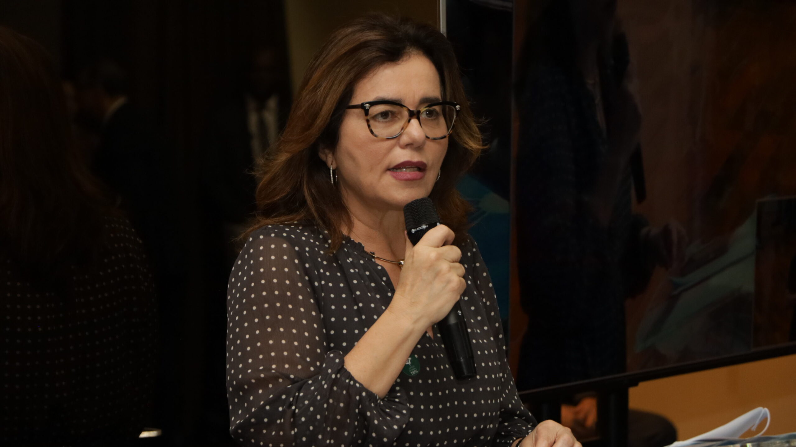 Uma mulher de meia-idade, vestindo uma blusa escura de bolinhas claras, falando em um microfone de mão. Ela usa óculos de aro claro e brincos de argola. O fundo está escuro e desfocado.