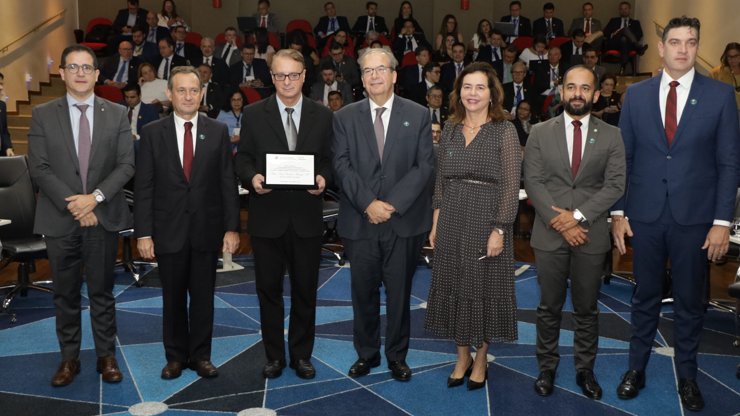 Uma foto de grupo com sete pessoas (seis homens e uma mulher) em trajes sociais formais, em pé na primeira fila. O terceiro homem da esquerda para a direita segura uma placa ou certificado emoldurado. A mulher veste um vestido longo estampado. Todos estão olhando para a câmera. Ao fundo, uma grande plateia de pessoas também em trajes formais está sentada em fileiras ascendentes. O chão é coberto por um tapete azul com padrão geométrico de linhas brancas.