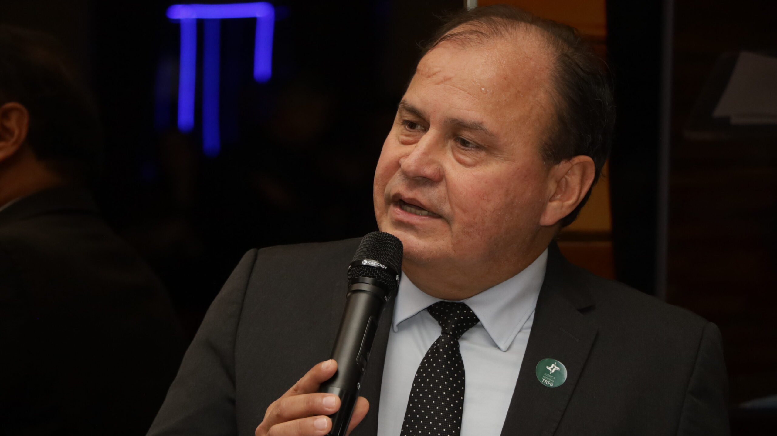 Um homem de meia-idade, vestindo terno escuro e gravata preta estampada, falando em um microfone de mão. Ele usa um broche na lapela direita. O fundo está desfocado e escuro, com reflexos de luzes azuis e brancas.