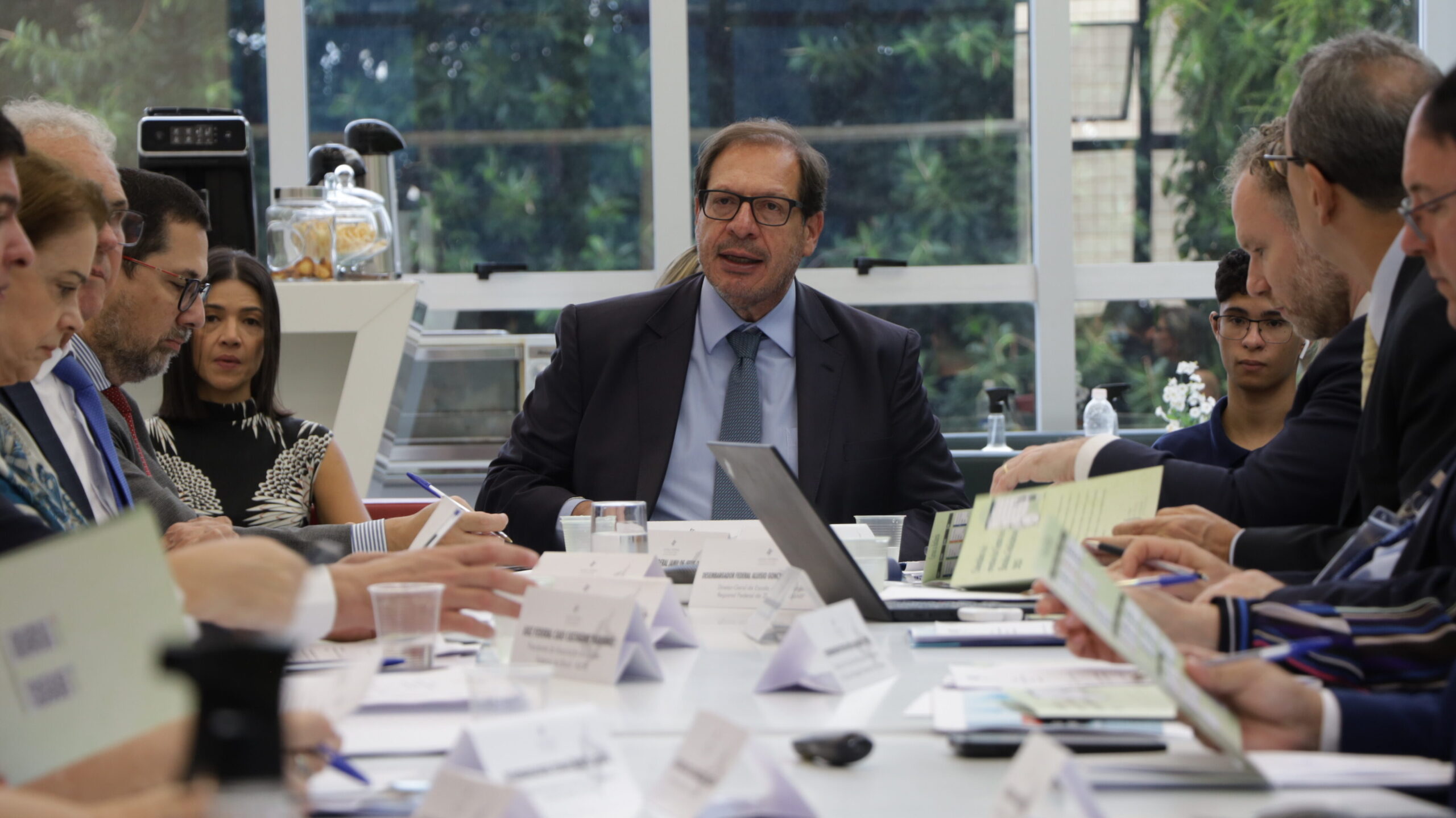 Foto em plano médio que retrata uma reunião formal em andamento, com múltiplas pessoas sentadas ao redor de uma mesa branca comprida.

O foco está no homem central, de terno escuro e gravata azul clara, que está falando e olhando para o grupo à sua direita. Ele usa óculos e gesticula levemente com as mãos sobre a mesa.

A mesa está cheia de materiais de reunião, cartões de identificação dobrados, copos e garrafas d'água. As pessoas ao redor estão atentas, algumas escrevendo ou segurando documentos.

O fundo é uma janela grande que permite ver a vegetação externa.