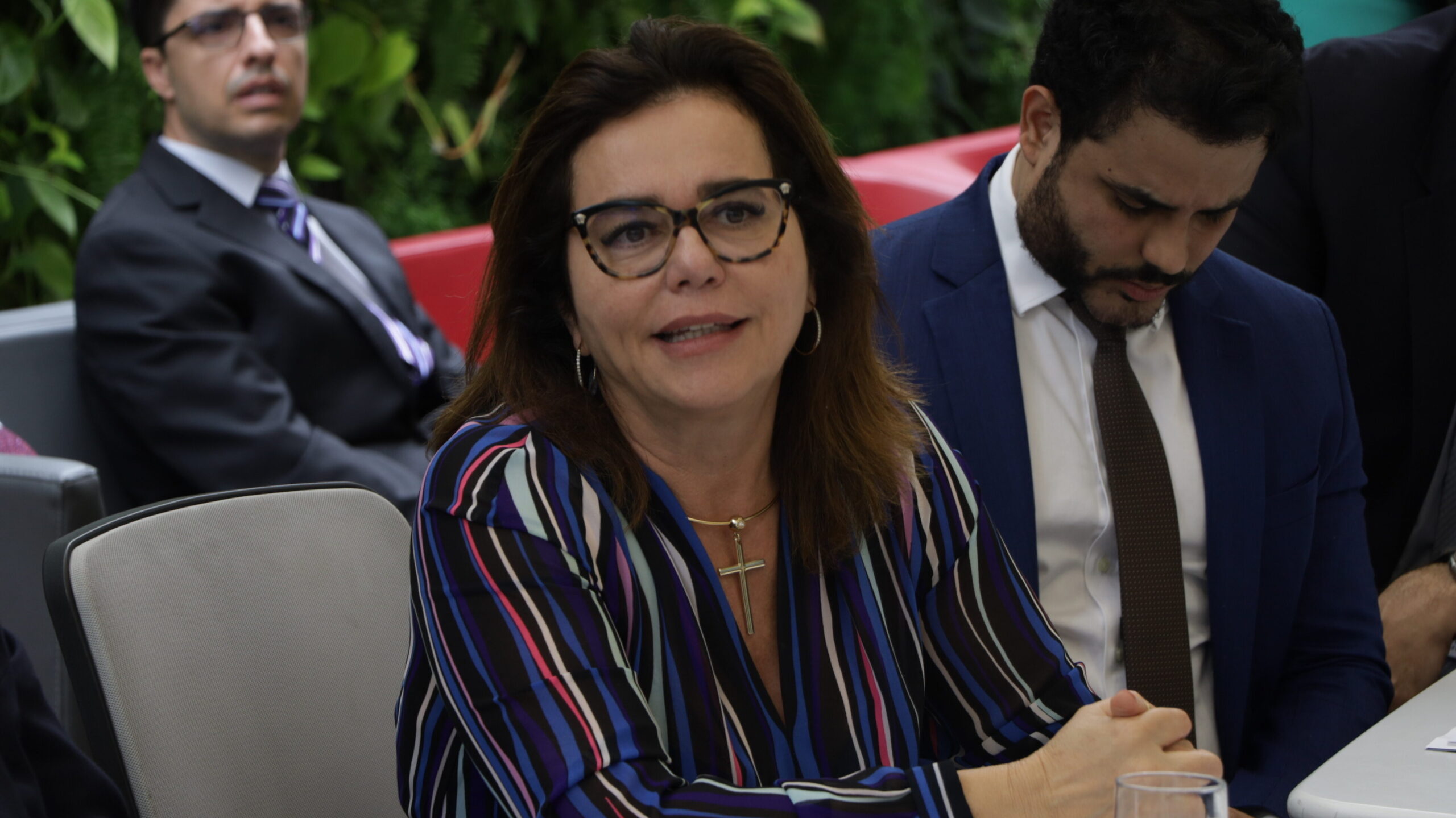 Foto em plano médio de uma mulher sentada em uma reunião ou evento.

Ela usa óculos, vestindo um vestido colorida (em tons de azul, roxo e branco) e um colar com pingente de cruz. Ela está com as mãos entrelaçadas à frente e olha ligeiramente para a sua esquerda, sorrindo de leve.

No fundo, parcialmente visíveis, estão dois homens sentados: um à esquerda de terno escuro, e outro à direita de terno azul-marinho. Atrás, é possível ver parte de uma parede de jardim vertical com folhagens verdes.