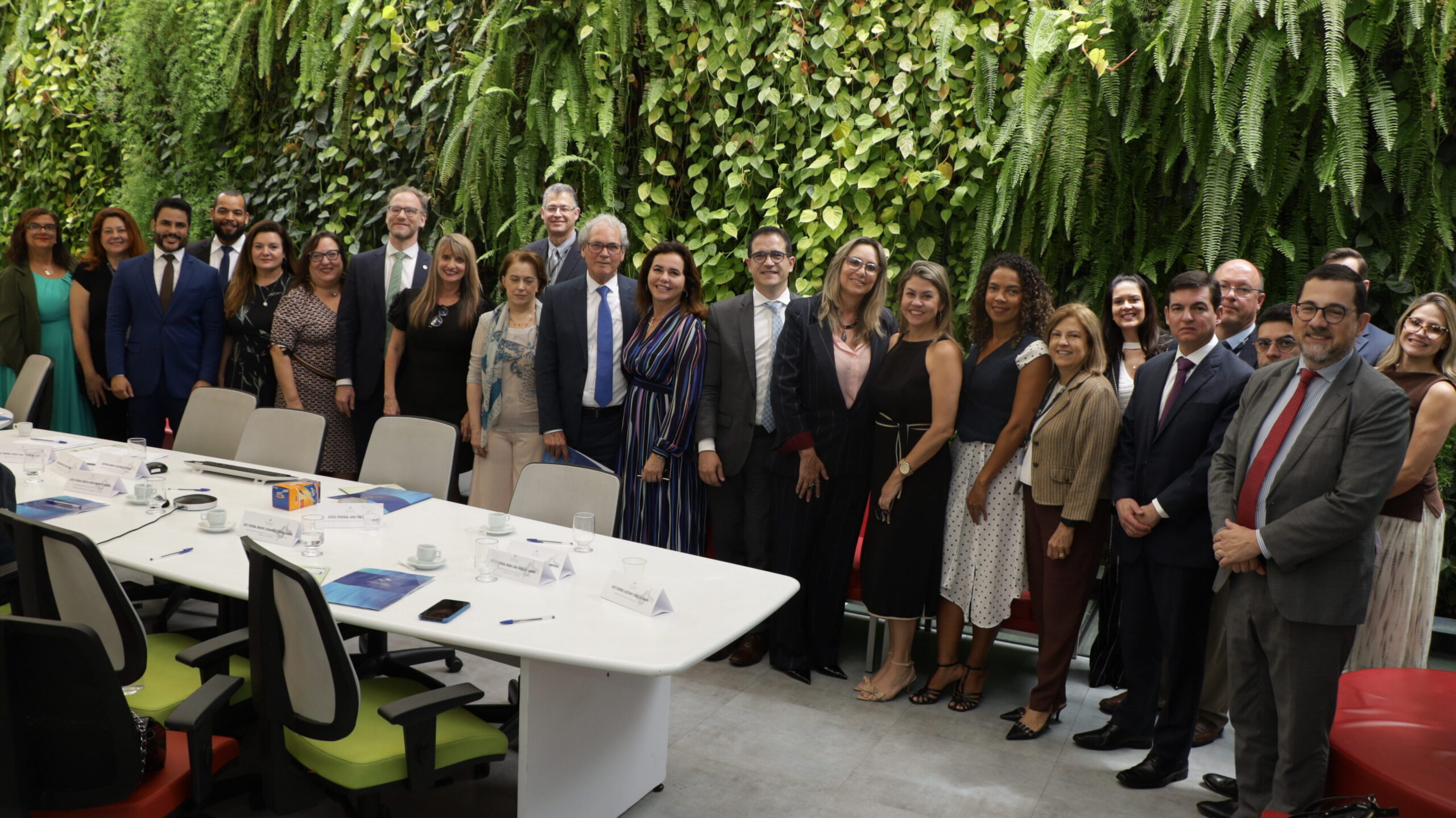Foto de grupo, de caráter institucional, em uma sala de reuniões moderna.

A foto reúne um grupo de pessoas (homens e mulheres) em pé, posicionadas atrás de uma grande mesa branca de reunião, que ocupa o primeiro plano. Todos estão vestidos com trajes sociais/formais de diversas cores.

O fundo é uma parede coberta por um jardim vertical de plantas e samambaias verdes.