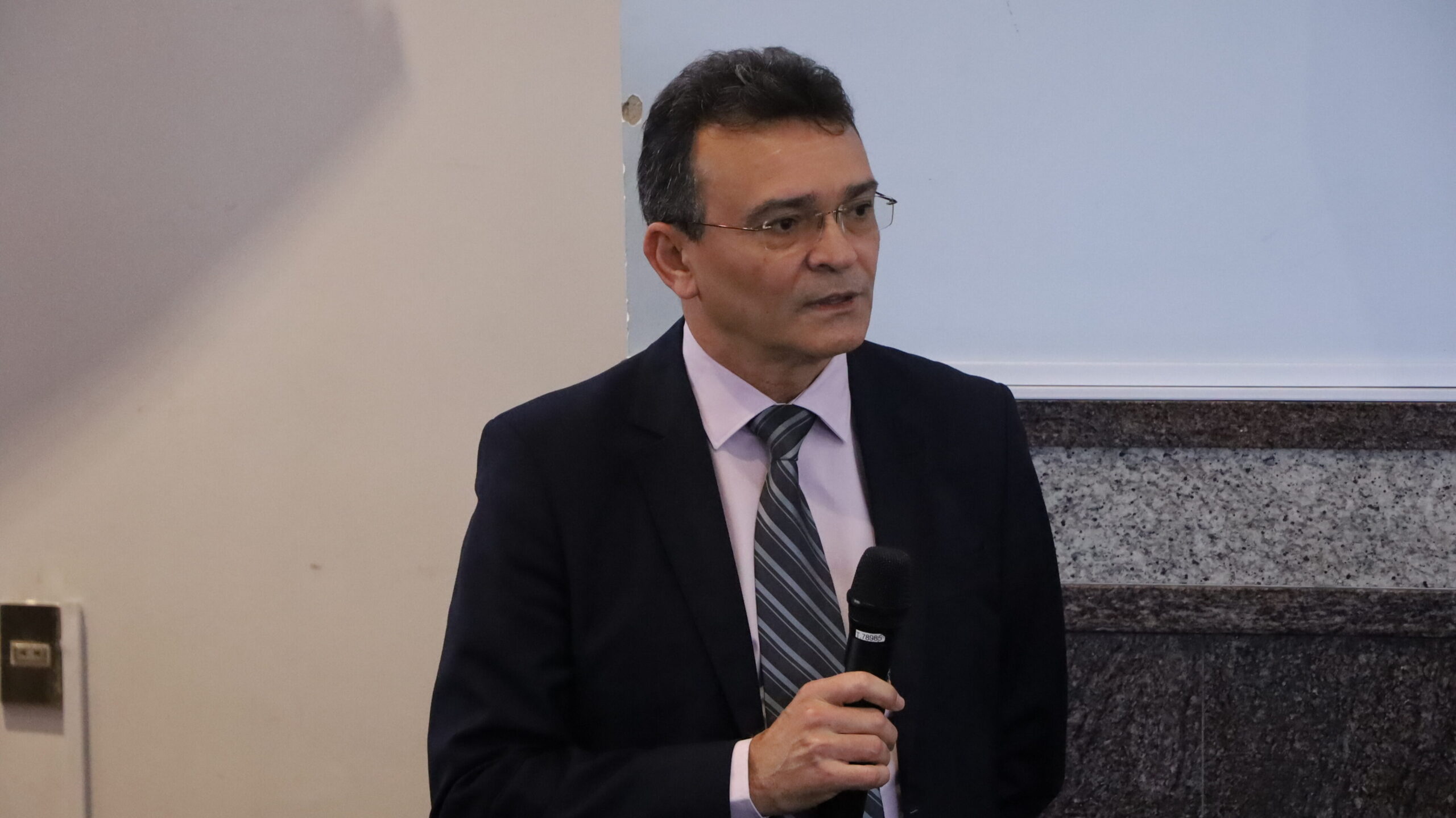 A imagem é uma fotografia de um homem em um ambiente interno. O homem está olhando para a frente e segurando um microfone preto com a mão direita. Ele veste um terno escuro, uma camisa social clara e uma gravata listrada em tons de cinza e preto.