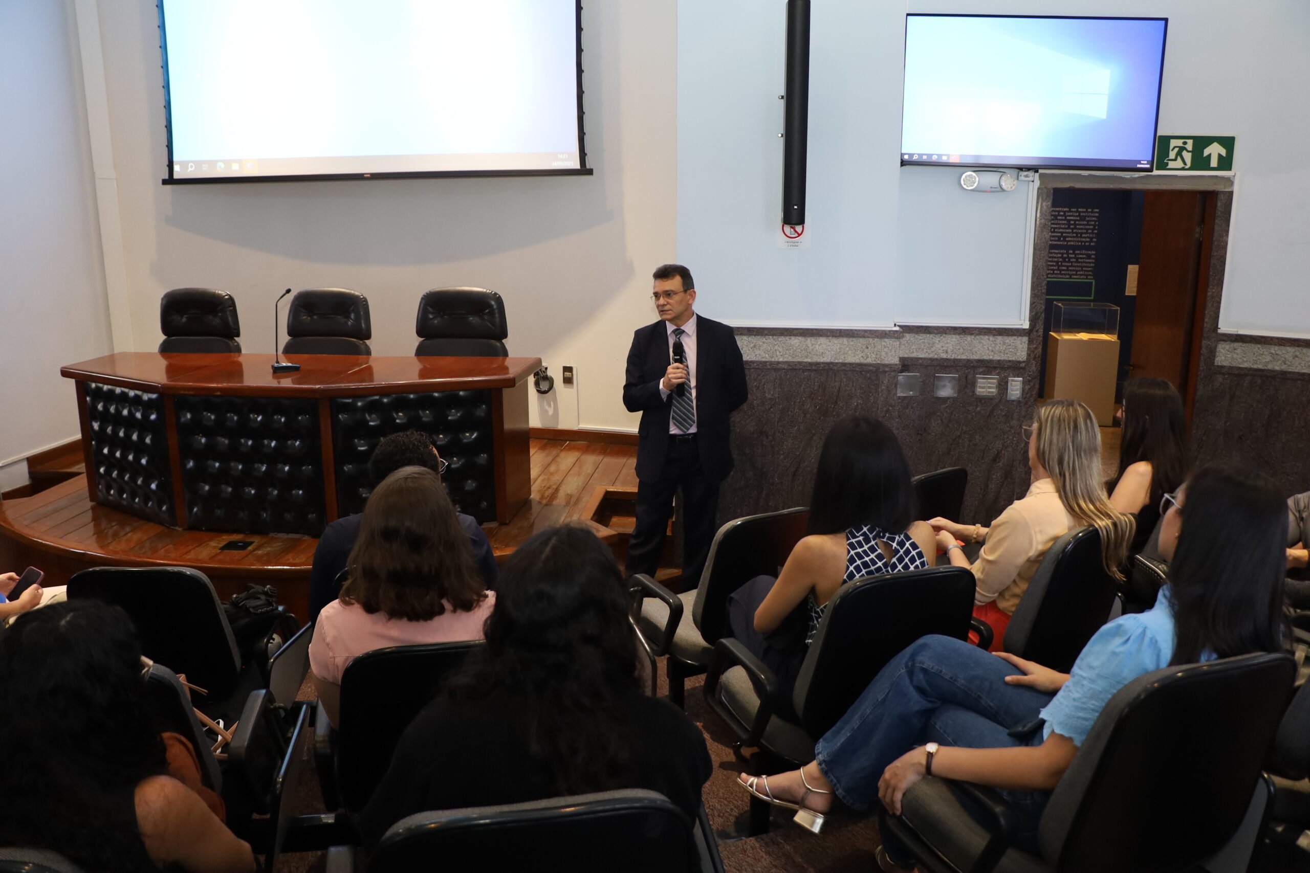 A imagem é uma fotografia de um evento ou apresentação em uma sala com assentos dispostos em auditório. Um homem em pé no centro, vestindo um terno escuro com gravata, está de frente para um grupo de pessoas sentadas e viradas de costas para a câmera. O homem segura um microfone na mão direita. Na área frontal e elevada, há uma mesa de madeira escura e curva com três cadeiras de couro pretas atrás dela. Na parede atrás do orador, há uma tela de projeção branca e, à direita, um monitor de TV ligado.