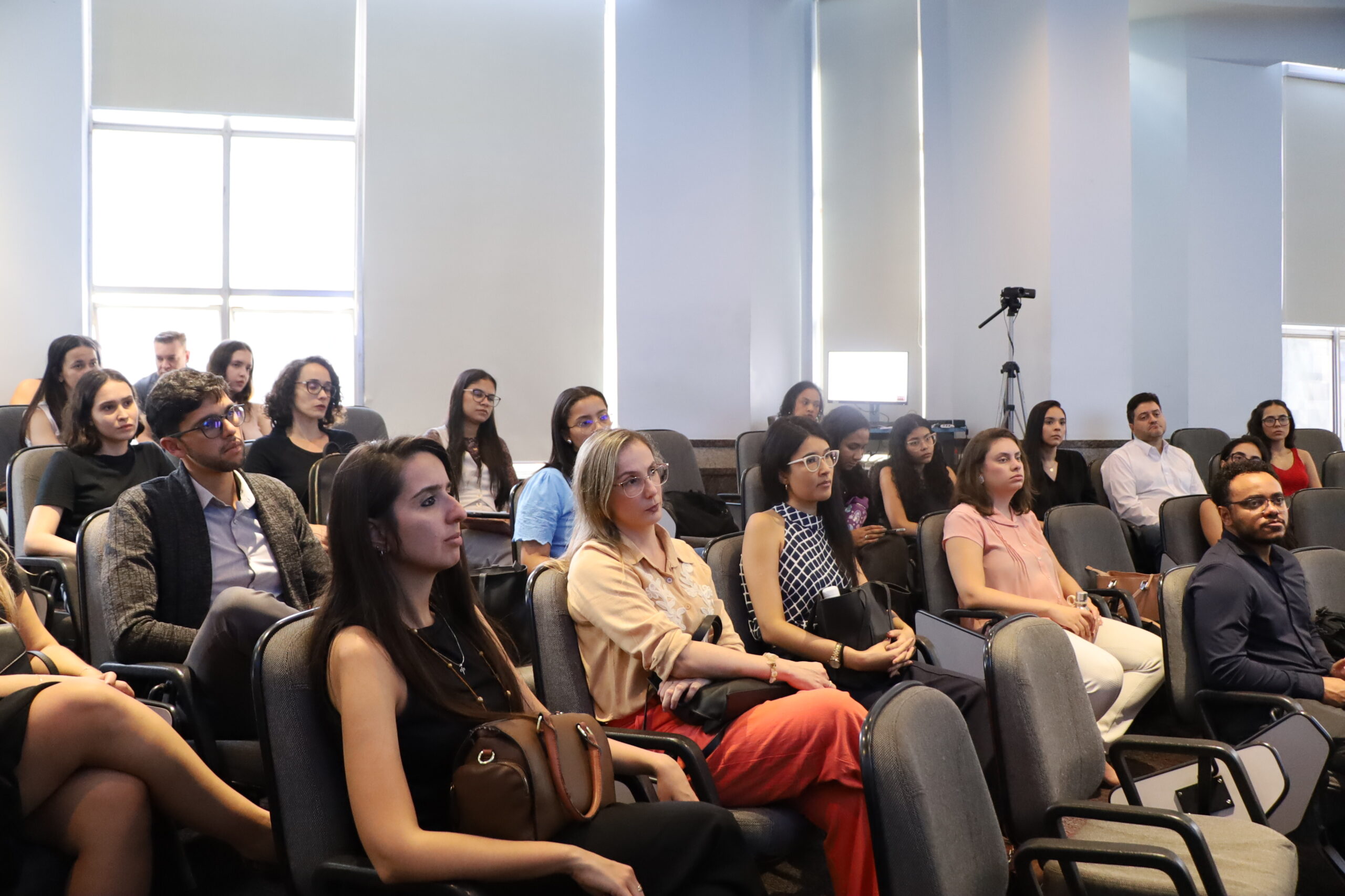 A imagem mostra um grupo de pessoas sentadas em uma sala de auditório. A maioria das pessoas está olhando na mesma direção. O ambiente interno é bem iluminado.