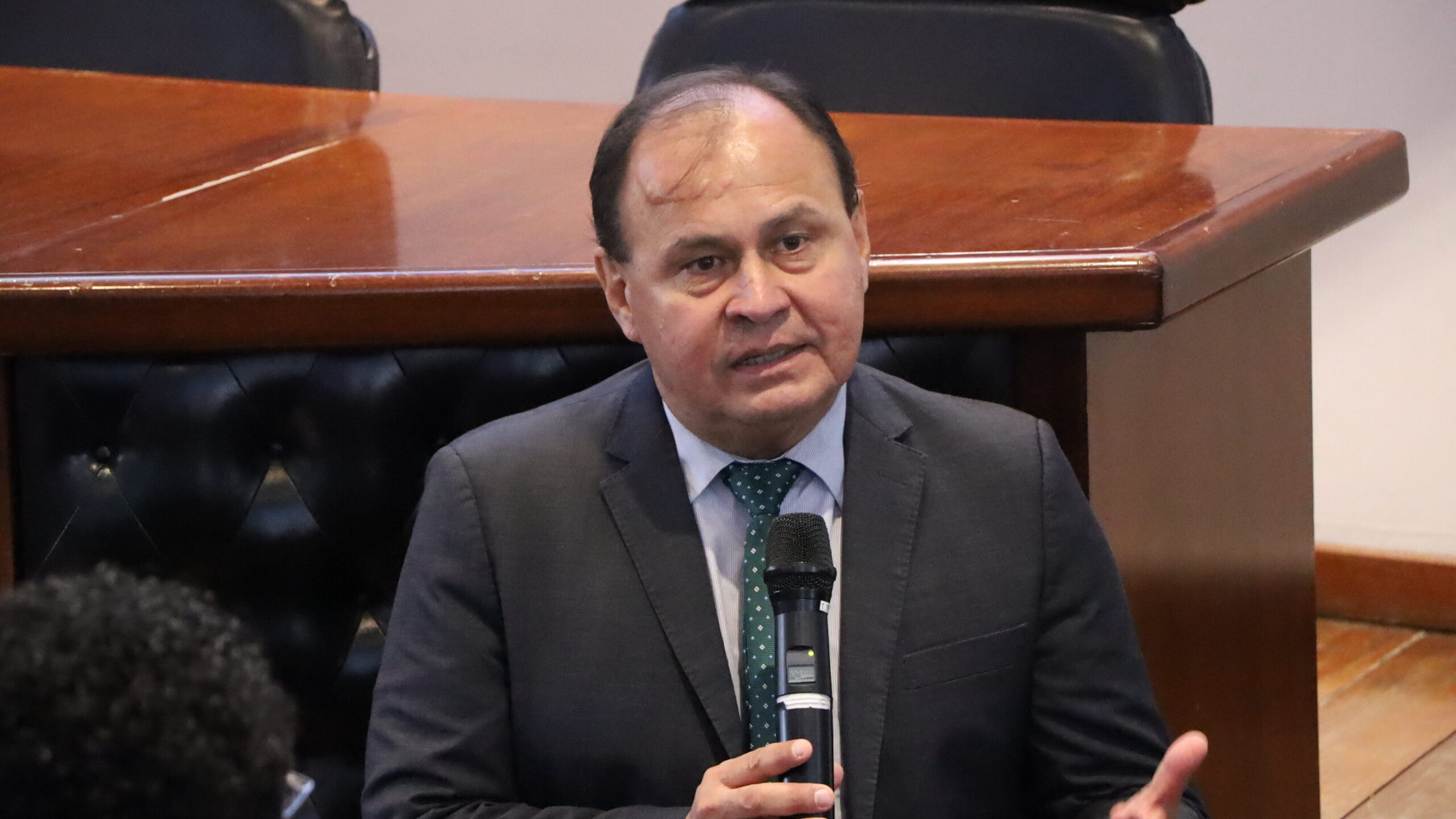 A imagem é uma fotografia em plano médio de um homem em um ambiente interno. O homem está olhando para a frente e segurando um microfone preto próximo ao peito com a mão direita, enquanto gesticula com a mão esquerda. Ele veste um terno escuro com uma gravata estampada em tons de verde. O homem está posicionado em frente a uma mesa de madeira escura.