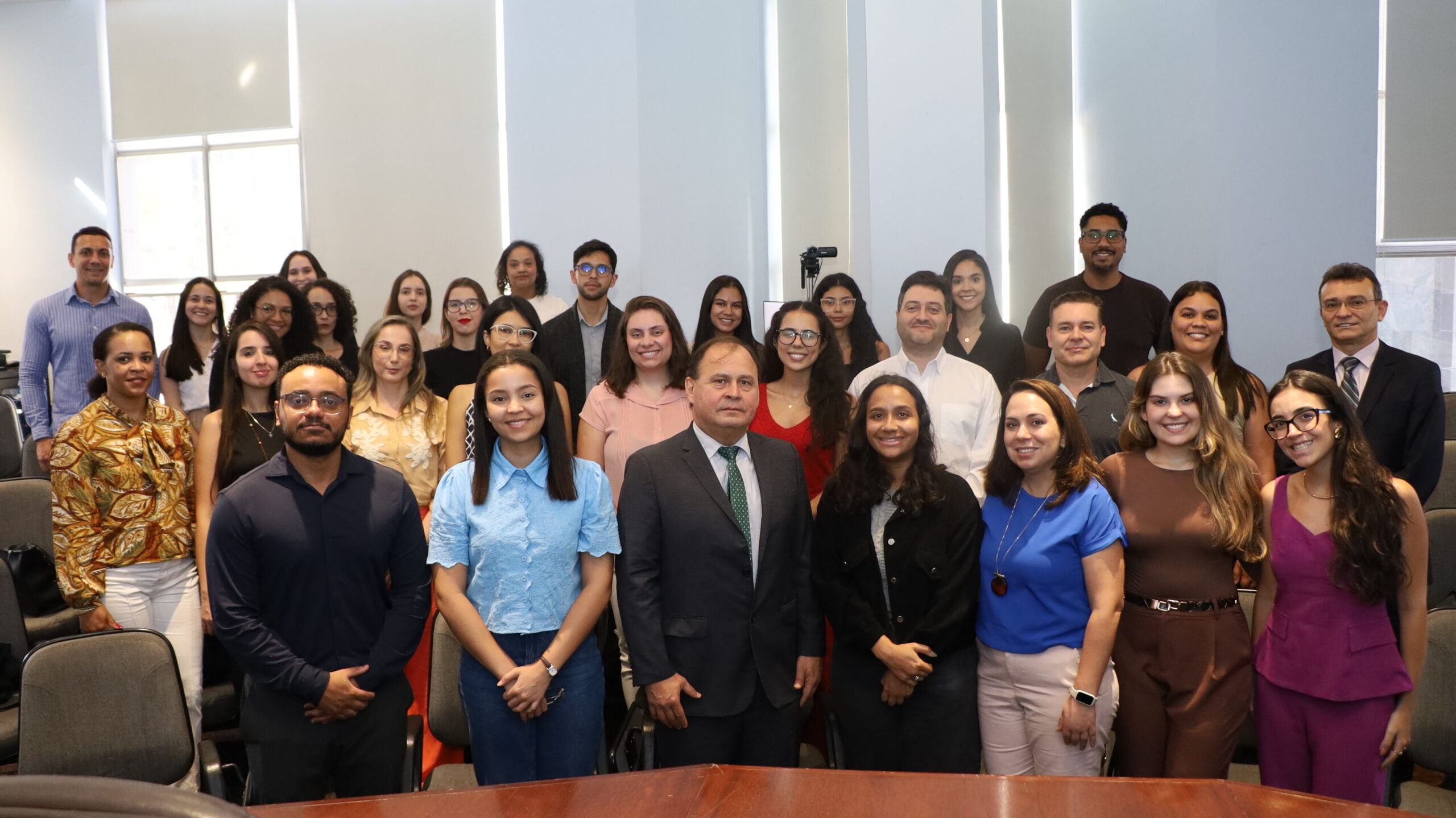 A imagem é uma fotografia de um grupo de pessoas, composto por homens e mulheres, de pé e posando para a câmera. O grupo está organizado em fileiras, atrás de uma mesa grande de madeira escura que ocupa o primeiro plano. Eles estão em um ambiente interno com paredes claras e grandes janelas visíveis no lado esquerdo, cobertas com persianas brancas. O grupo apresenta uma variedade de vestimentas, predominando trajes casuais e sociais.