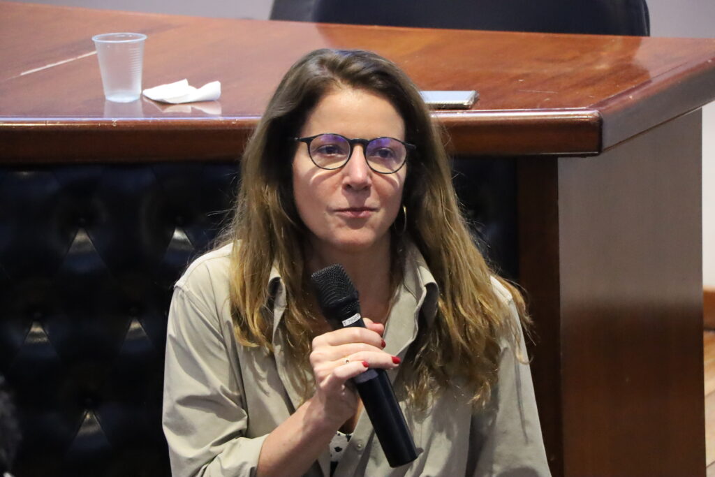 A imagem é uma fotografia de uma mulher em um ambiente interno. A mulher está olhando para a frente e segurando um microfone preto com a mão direita, próxima ao rosto. Ela usa óculos de grau e uma camisa de cor cáqui ou bege com as mangas dobradas. A mulher está posicionada em frente a uma mesa de madeira escura.