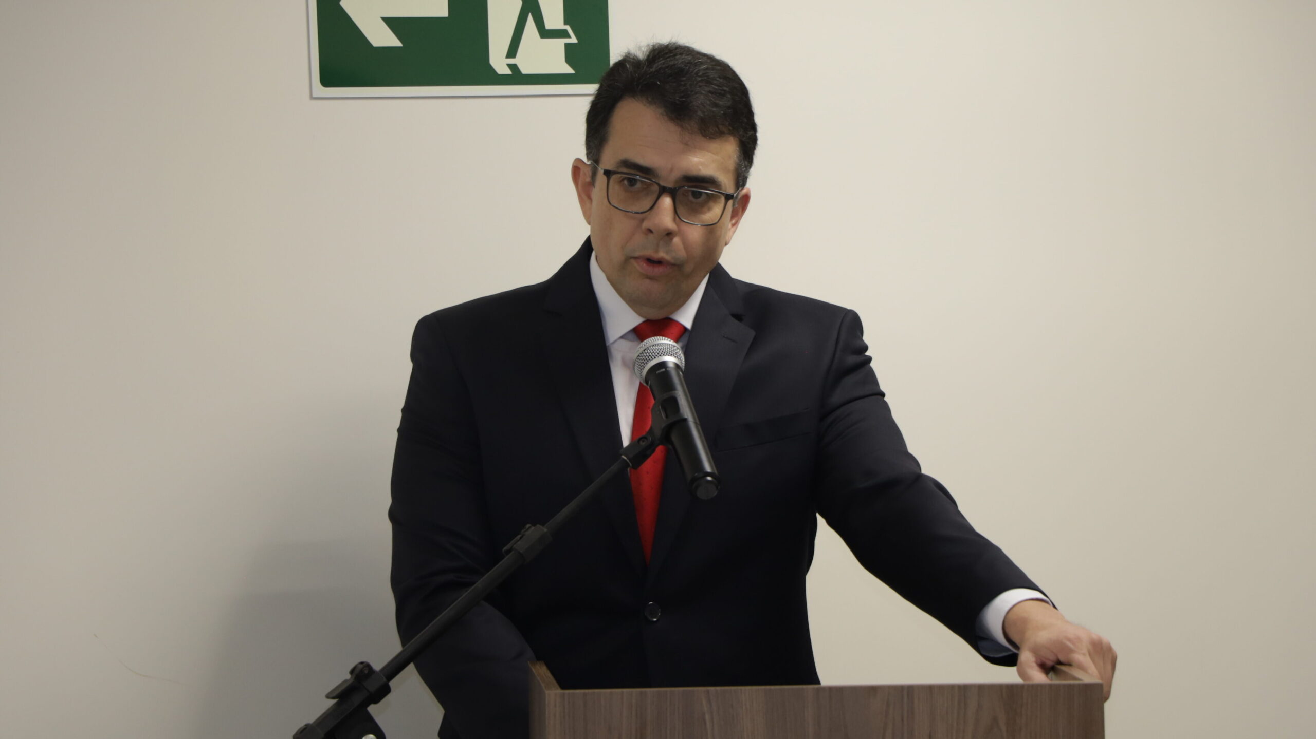 Um homem de óculos, vestindo um terno escuro e uma gravata vermelha, está falando a um microfone fixo em um púlpito ou pódio de madeira escura. A foto é um plano médio do orador em um ambiente interno formal, com um fundo de parede branca.