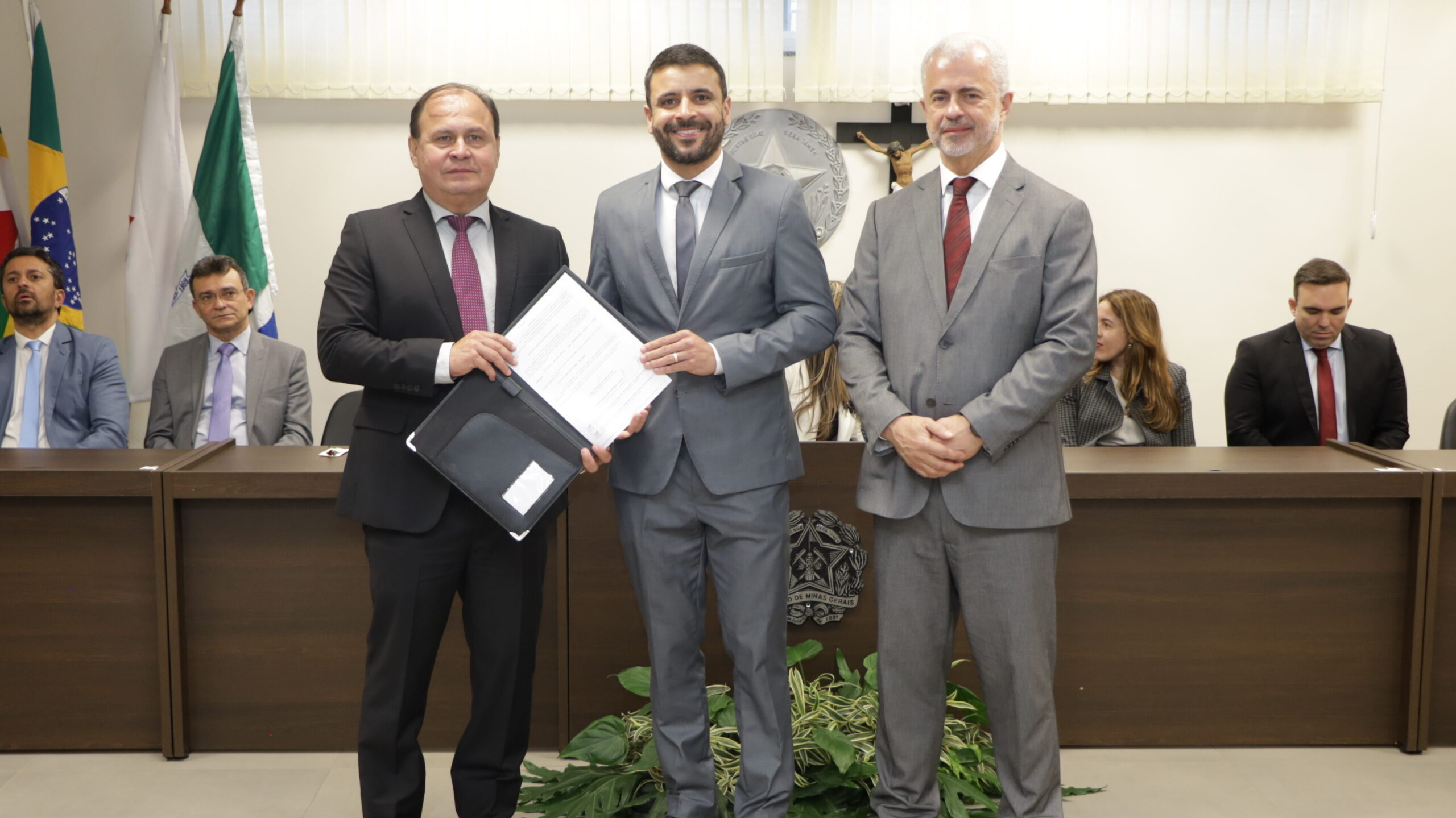 Três homens em trajes formais estão de pé no centro de uma sala, participando de uma assinatura de documentos.

O homem da esquerda, de terno escuro, segura uma pasta preta com documentos abertos. O homem no centro, de terno cinza, está com a mão sobre os documentos. O homem da direita, de terno cinza, está com as mãos postas à frente.

No fundo, há uma mesa escura onde outros participantes estão sentados. Acima, algumas bandeiras  são visíveis.