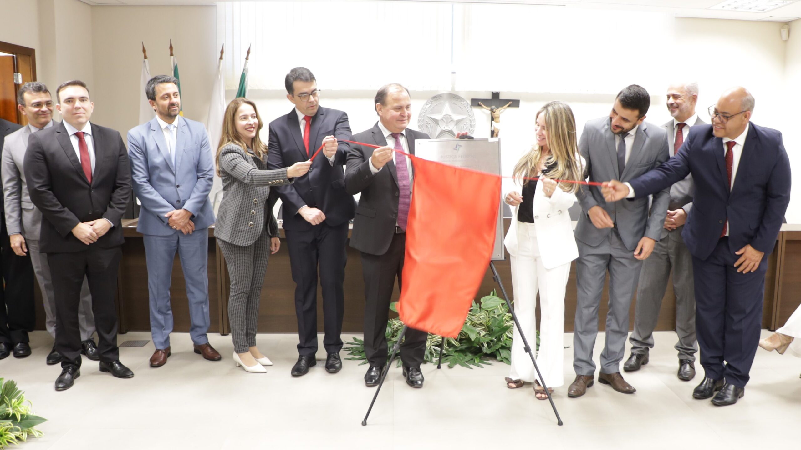 Um grupo de dez pessoas (oito homens e duas mulheres) em trajes sociais está participando de uma cerimônia de descerramento de uma placa.

O homem no centro está retirando um tecido vermelho que cobre a placa, enquanto um homem e uma mulher, cada um de um lado, auxiliam segurando as fitas de descerramento. O grupo está em pé em um ambiente formal com bandeiras ao fundo à esquerda, uma cruz na parede e folhagens decorativas no chão.