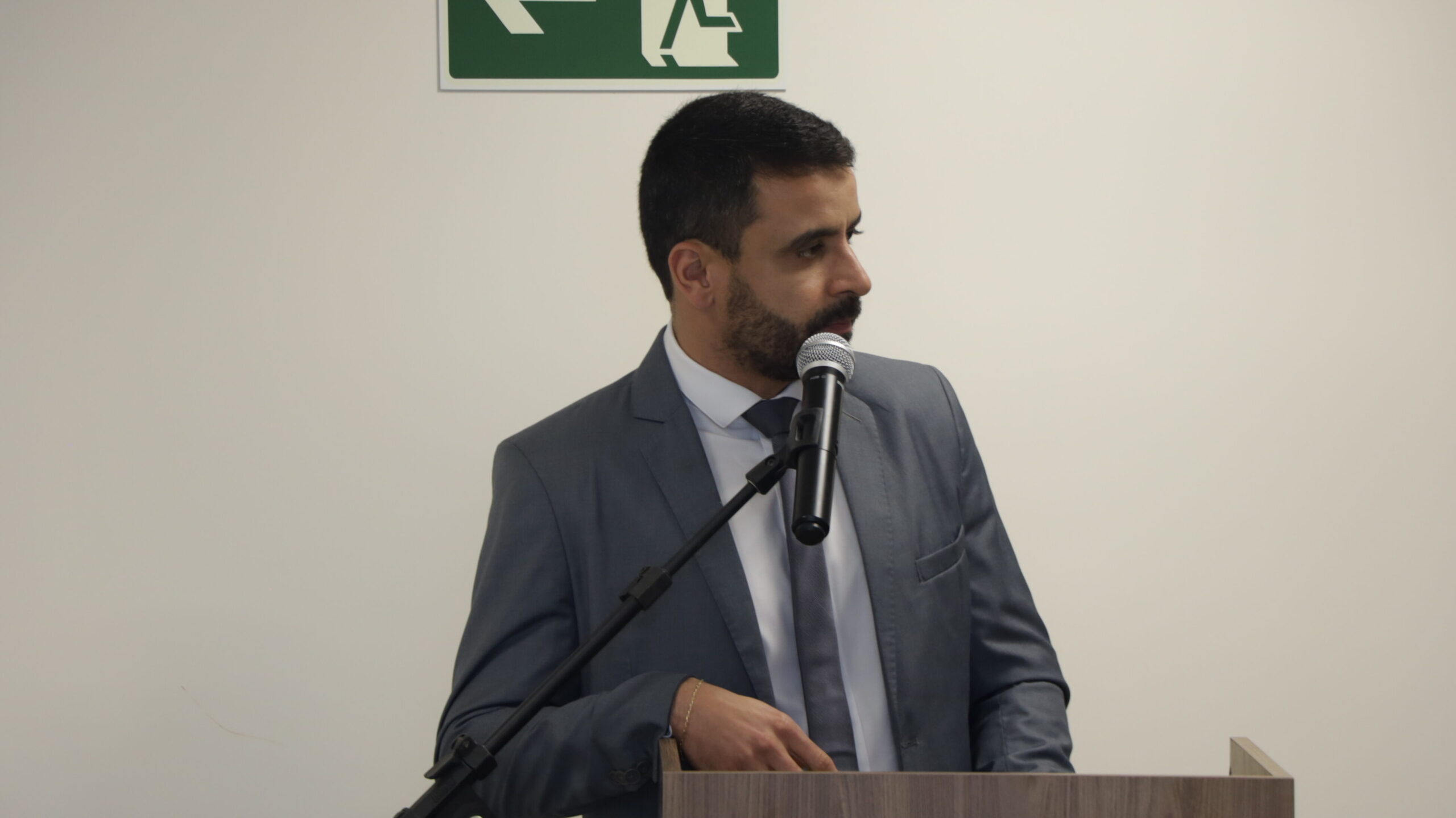 Um homem de barba, vestindo um terno cinza e gravata, está falando a um microfone fixo em um púlpito ou pódio de madeira. Ele olha para o lado direito. A foto é um plano médio do orador em um ambiente interno formal, com um fundo de parede branca.