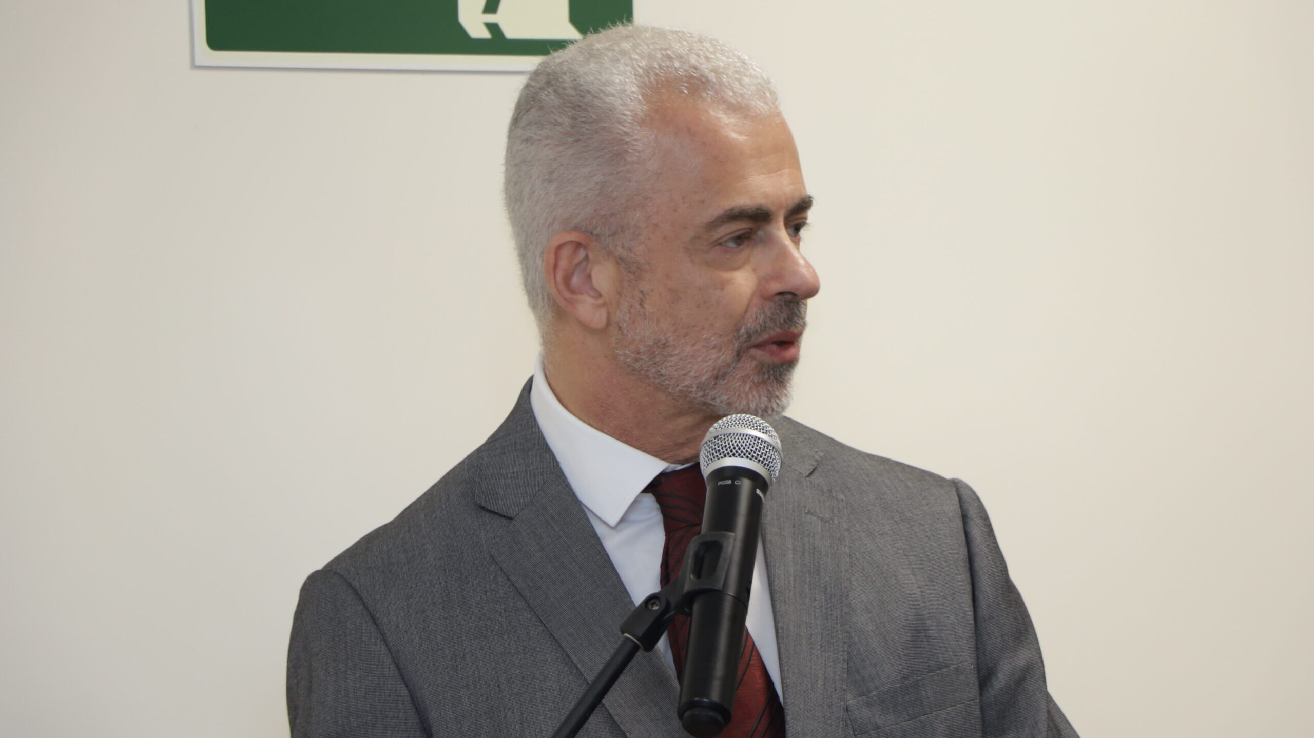 Um homem de cabelos grisalhos, vestindo um terno cinza e uma gravata bordô estampada, está falando a um microfone. A foto é um plano médio do orador em um ambiente interno formal, com um fundo de parede branca e lisa.