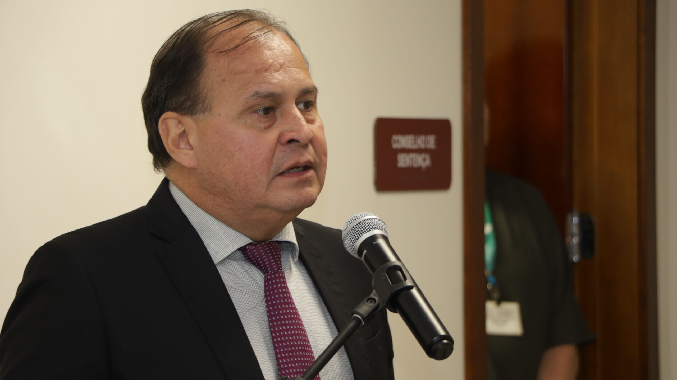 Um homem de meia-idade, vestindo um terno escuro e uma gravata bordô estampada, está falando a um microfone que está em primeiro plano.

A foto é um plano médio do orador em um ambiente interno, com uma porta escura e uma placa marrom na parede que diz "CONSELHO DE SENTENÇA" ao fundo. Uma pessoa é parcialmente visível atrás da porta.