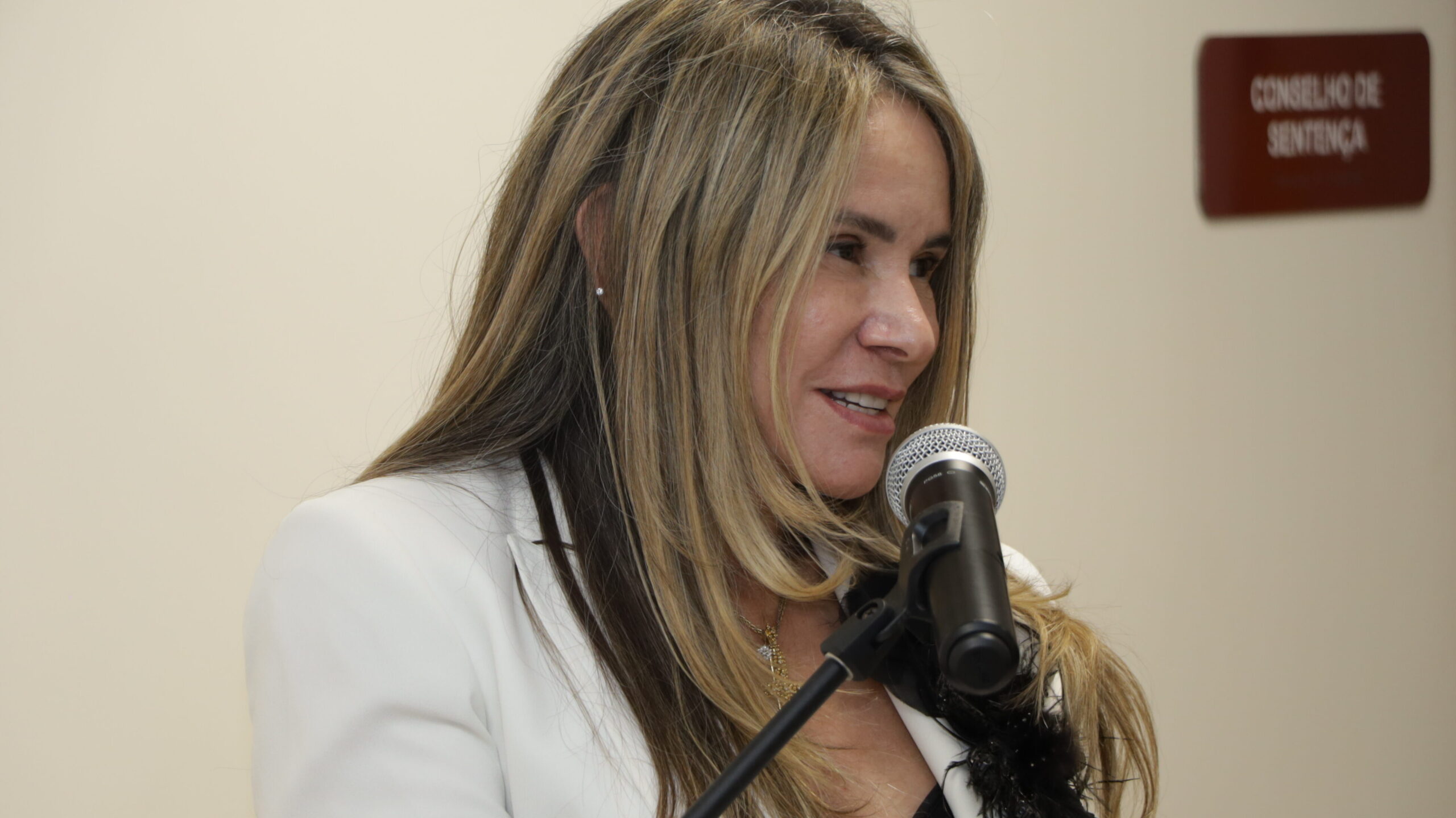 Uma mulher de cabelos loiros e compridos está falando a um microfone. Ela veste um blazer branco sobre uma roupa preta. A foto é um plano próximo da oradora em um ambiente interno formal, com um fundo de parede bege e uma placa marrom com o texto "CONSELHO DE SENTENÇA" no canto superior direito.