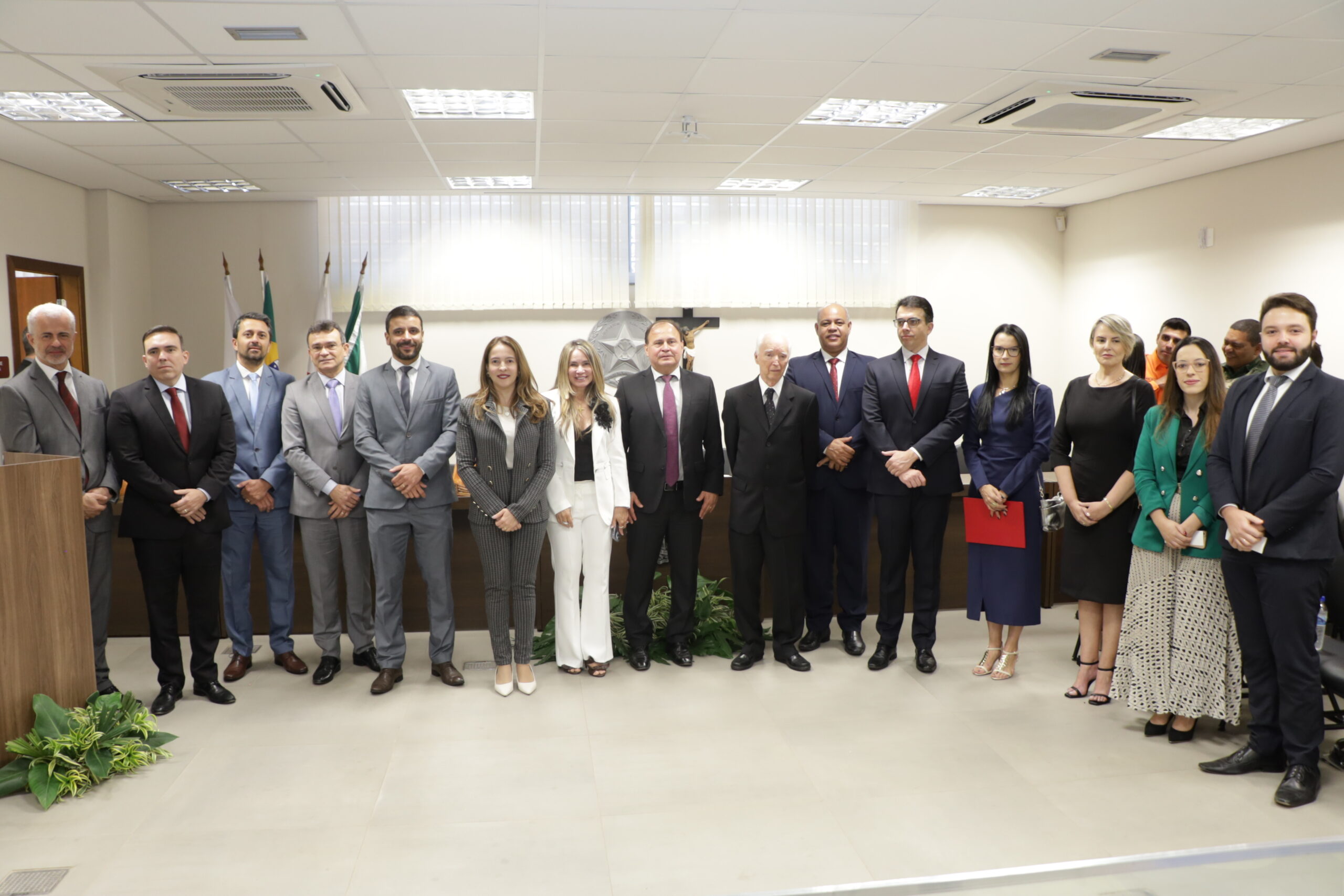 Um grupo de pessoas em trajes sociais está posando em pé em uma sala de conferências. Os homens vestem ternos de cores variadas e as mulheres usam vestidos ou terninhos. O ambiente é formal, com bandeiras na lateral e um crucifixo na parede central. Há arranjos de folhagens verdes no chão.
