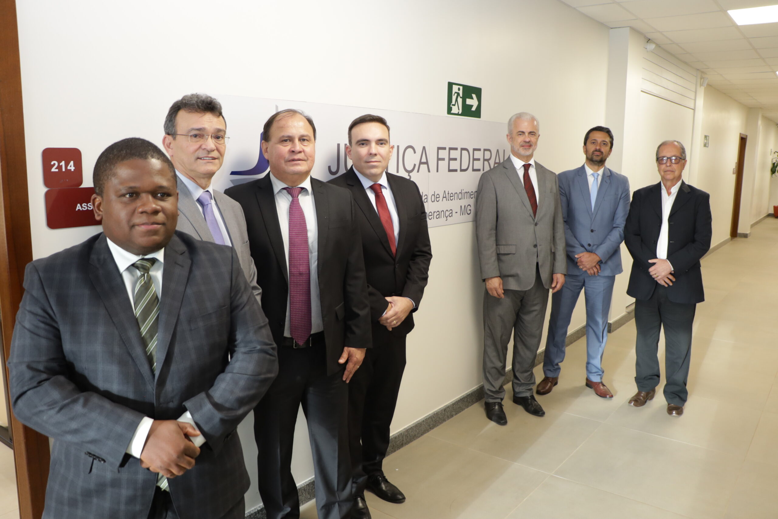 Um grupo de sete homens de terno posando em um corredor de escritório bem iluminado, ao lado de uma parede com um texto que inclui "JUSTIÇA FEDERAL".