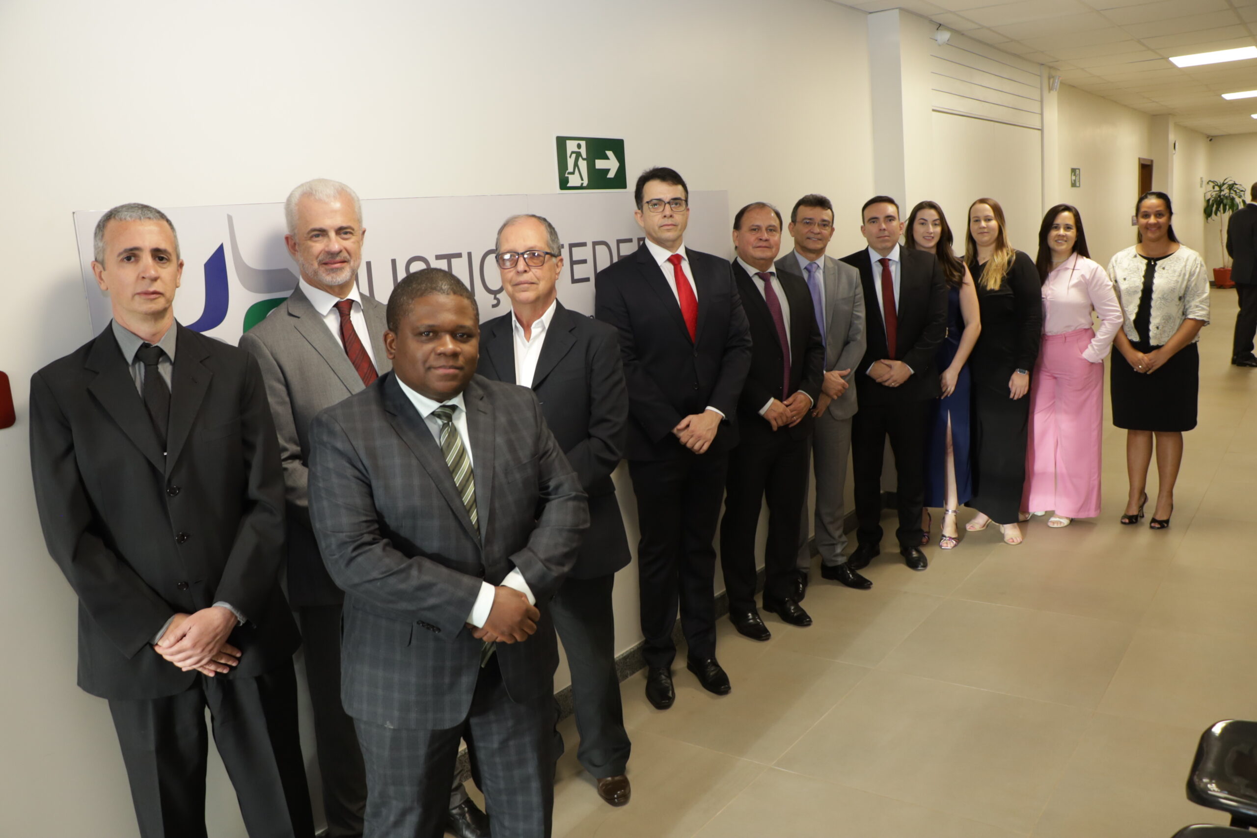 Um grupo de pessoas, sendo a maioria homens de terno e algumas mulheres de vestido social, posando em fila em um corredor de escritório ao lado de um letreiro com o texto "JUSTIÇA FEDERAL".