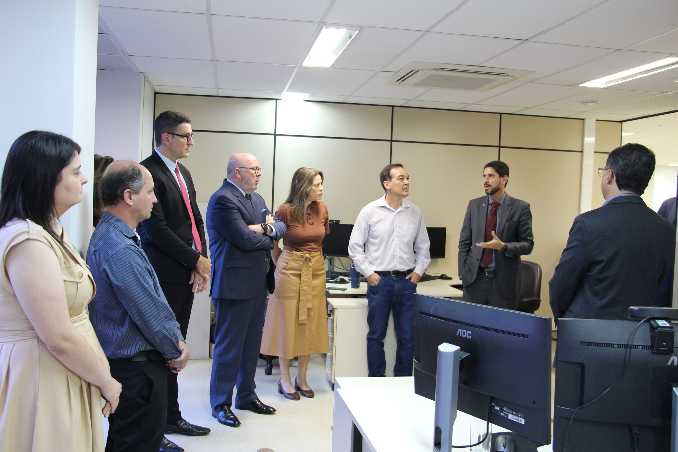 Em uma sala de escritório, um grupo de pessoas está reunido, com um homem de blazer e barba, no centro, falando e gesticulando. Os demais membros do grupo, todos em trajes sociais, estão ao seu redor, ouvindo atentamente. Na sala, há mesas de trabalho com computadores.