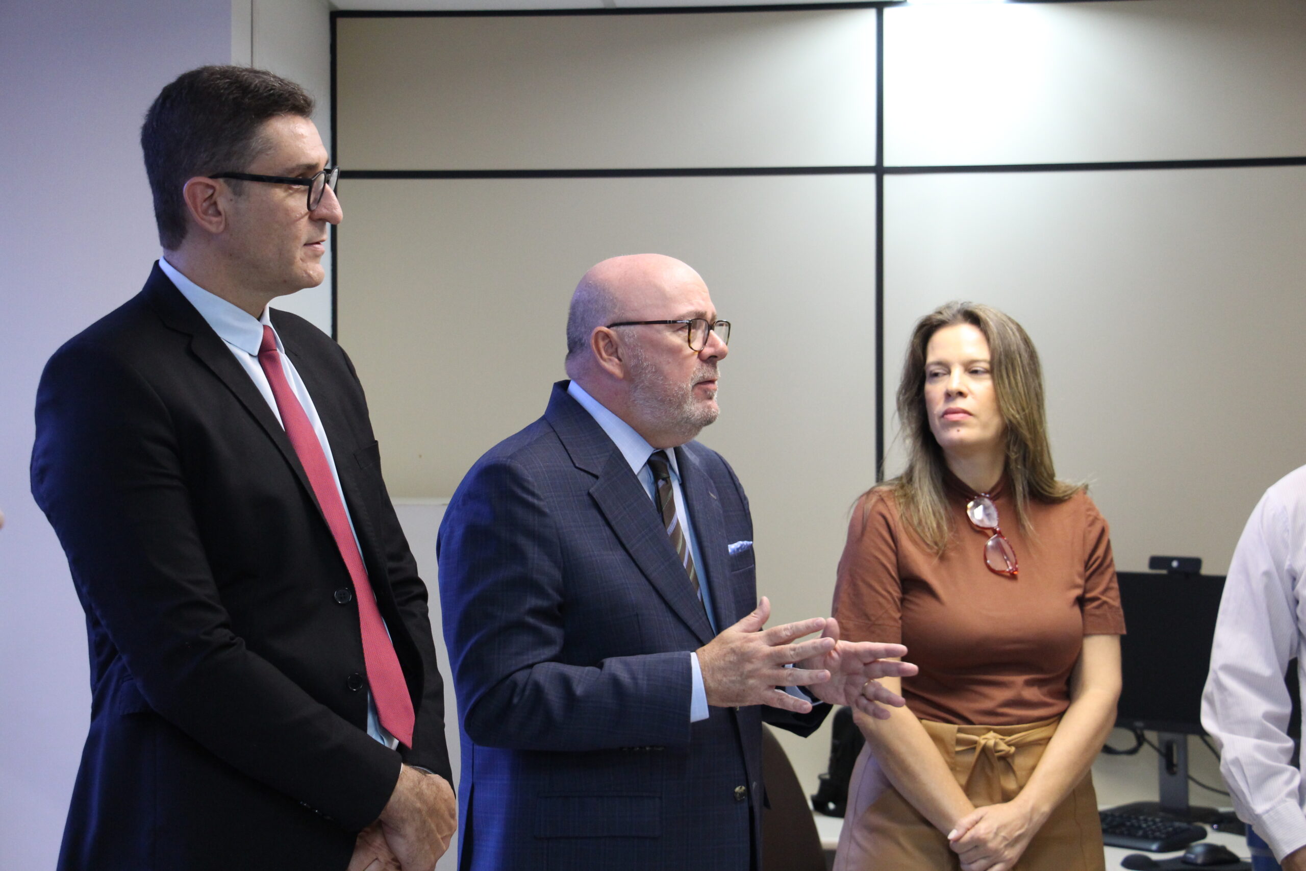 Três pessoas estão em pé em uma sala de escritório, sendo dois homens e uma mulher. No centro, um homem calvo de barba e óculos está com as mãos gesticulando enquanto fala. Um homem mais alto, de terno e gravata vermelha, está à esquerda, ouvindo atentamente, e uma mulher com uma blusa marrom e saia bege está à direita.