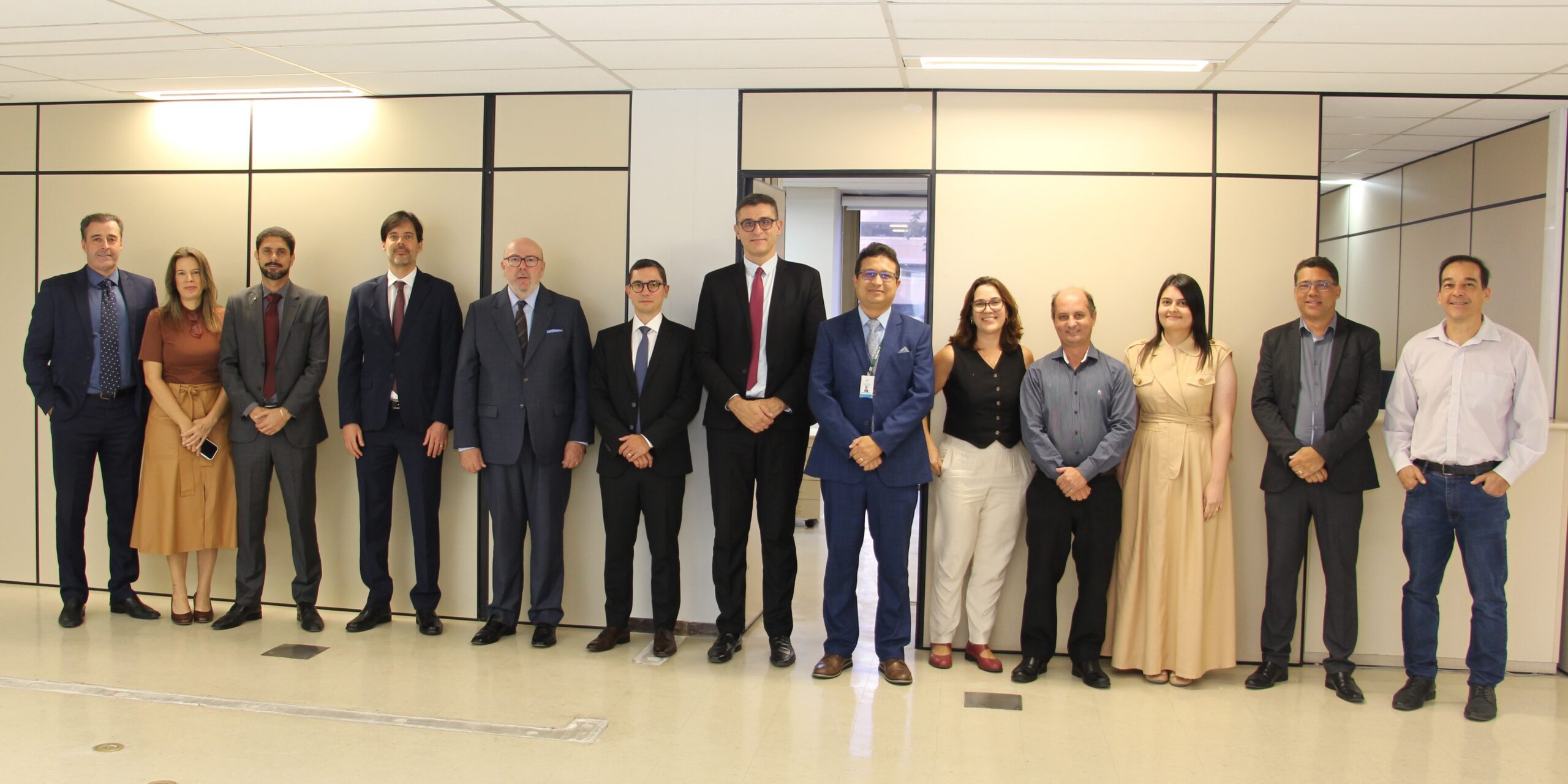Em pé, em uma sala, um grupo de treze pessoas, sendo dez homens e três mulheres, está posando para uma foto. Todos estão vestindo trajes sociais. No fundo, há uma porta aberta e divisórias de escritórios.