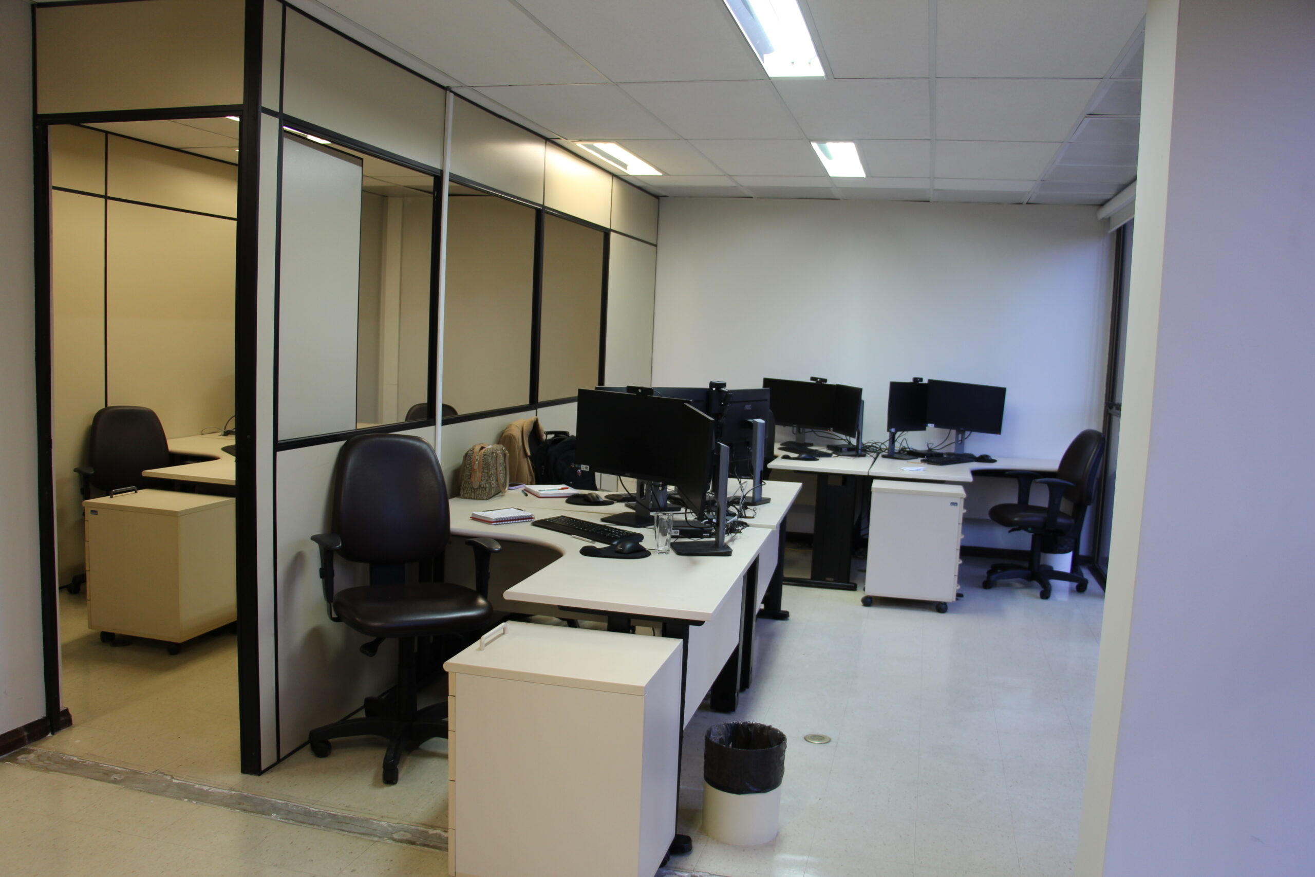 A imagem mostra uma sala de escritório com várias mesas dispostas em L, equipadas com monitores de computador e cadeiras de escritório. Há armários baixos com rodinhas ao lado das mesas. O espaço é dividido por paredes divisórias brancas e há um espelho em um dos lados. O chão é claro e há luminárias no teto.