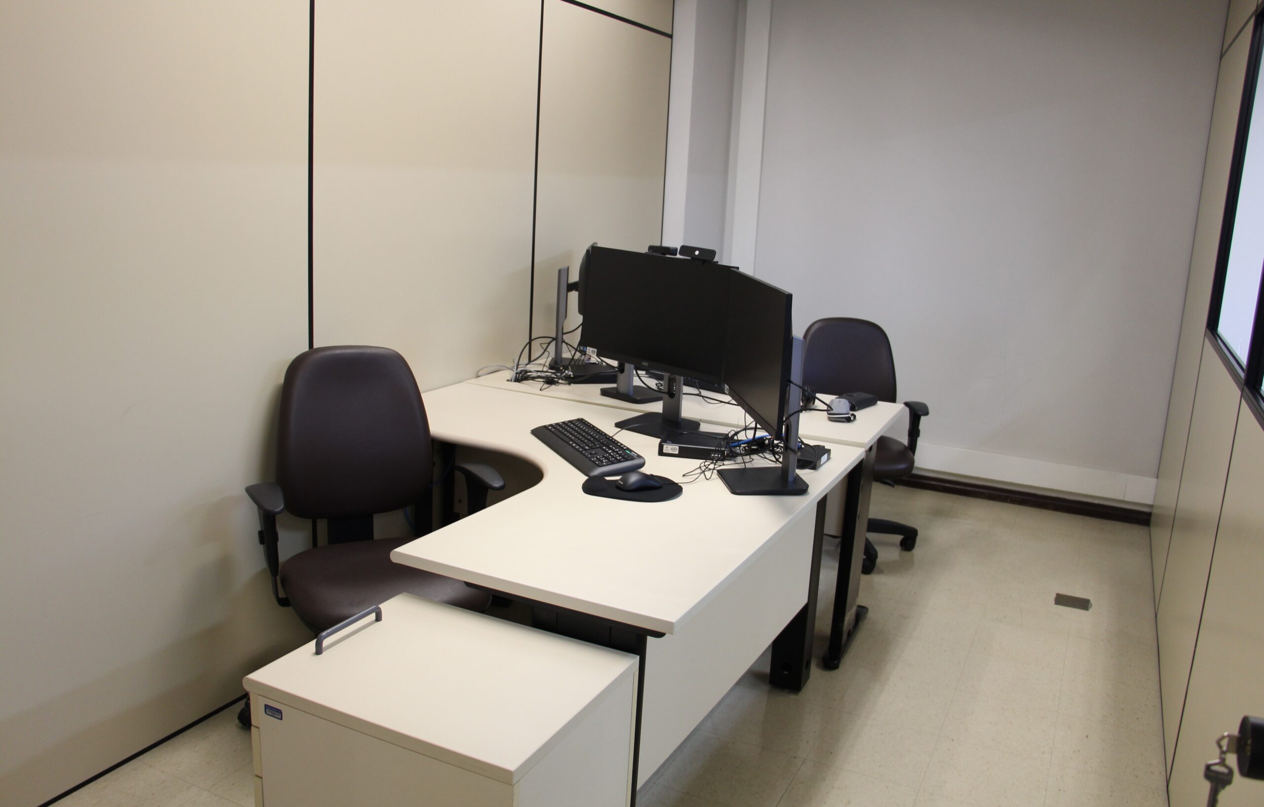 A imagem mostra uma estação de trabalho de escritório, com uma mesa de canto em formato de L e duas cadeiras. Há dois monitores e teclados sobre a mesa, além de um armário baixo com rodinhas. As paredes são brancas com divisórias e o chão é claro.