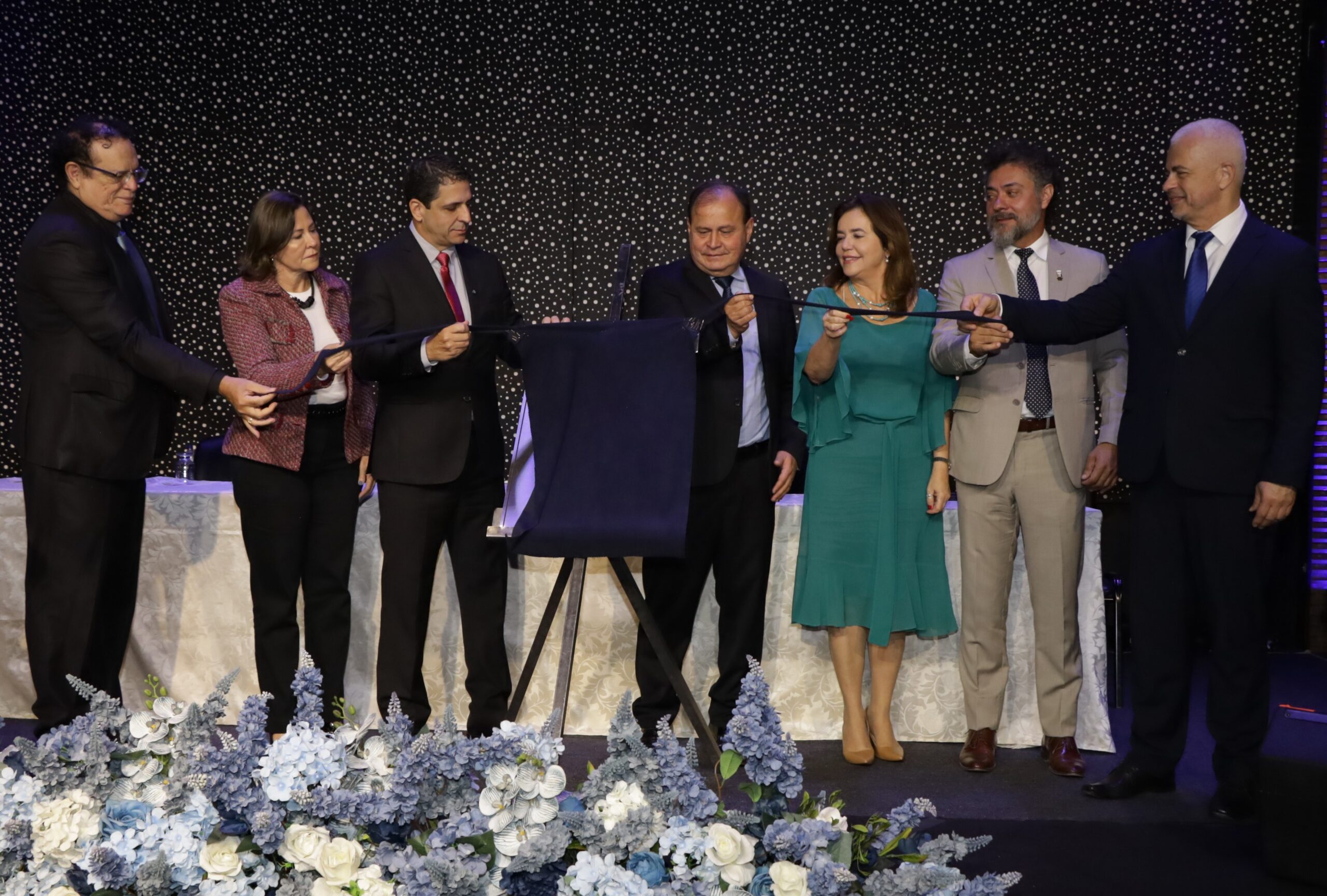 Sete pessoas, incluindo cinco homens e duas mulheres, estão em um palco. Eles participam de uma cerimônia, com algumas delas segurando uma fita preta e outras, um tecido azul-escuro sobre uma placa. Há um grande arranjo floral azul e branco no chão, à frente do palco.