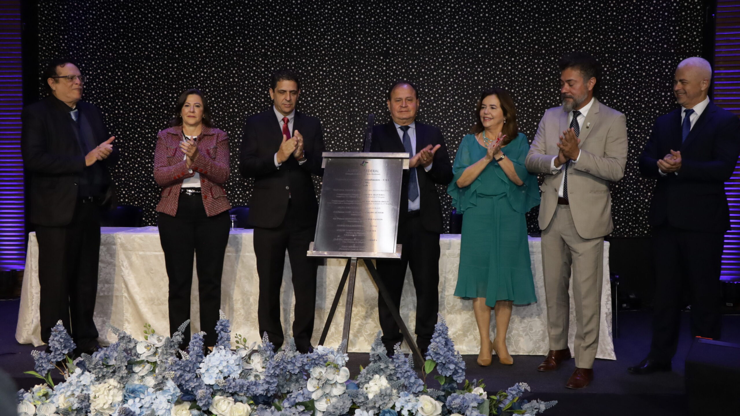 A imagem é de uma cerimônia onde seis pessoas estão em frente a uma placa comemorativa coberta, enquanto as duas pessoas nas extremidades, um homem e uma mulher, aplaudem.

No centro, um grupo de quatro pessoas, incluindo dois homens e duas mulheres, estão de pé, lado a lado, com o homem do meio batendo palmas. A mulher ao seu lado veste um vestido verde-água e o homem da ponta direita veste um terno cinza-claro. O palco é formal, com um fundo preto texturizado e uma mesa com toalha branca. Na frente do palco, há arranjos florais em tons de azul e branco.