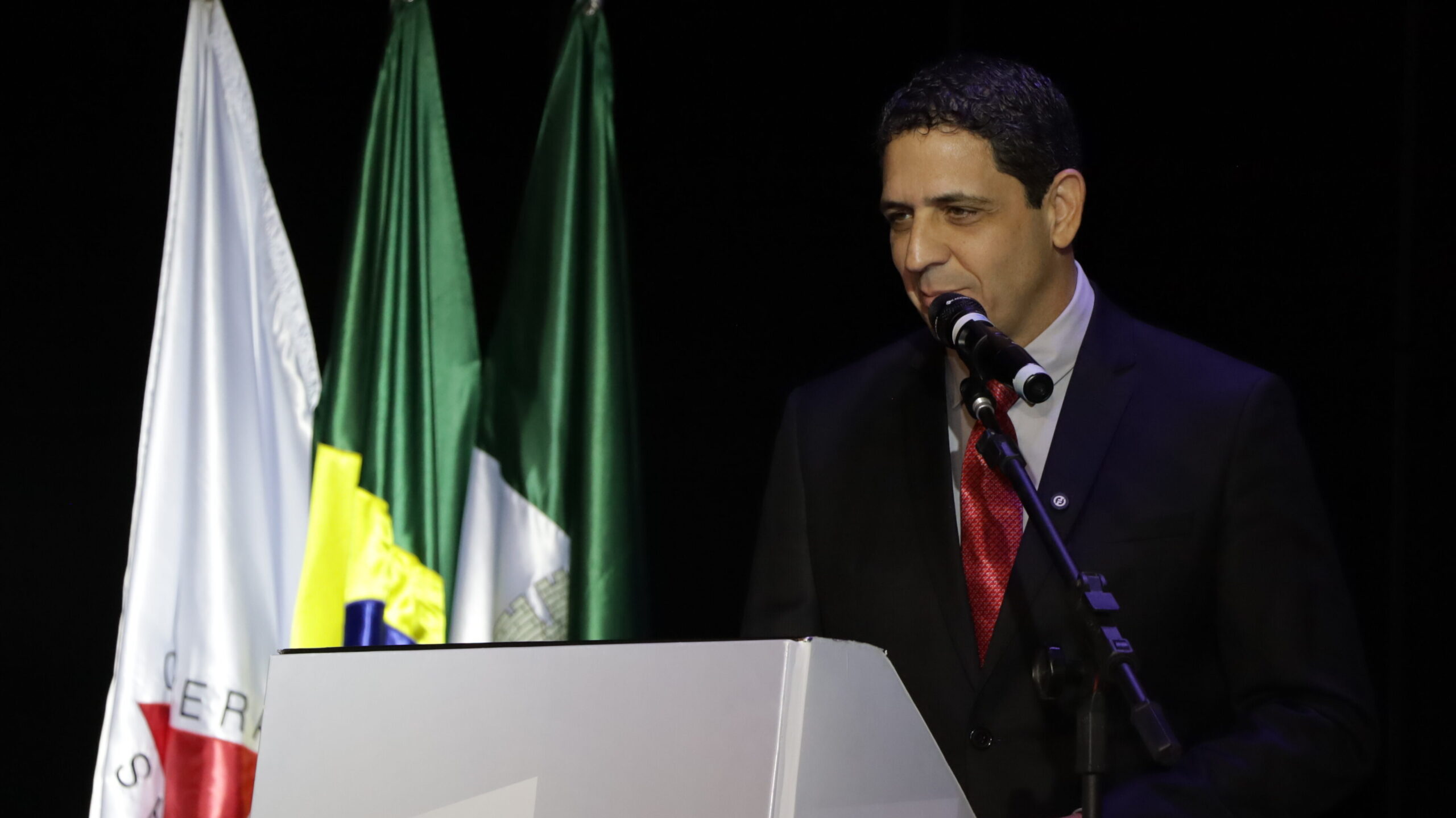 Um homem de terno preto, camisa branca e gravata vermelha está em um pódio, falando em um microfone. Atrás dele, três bandeiras são visíveis.